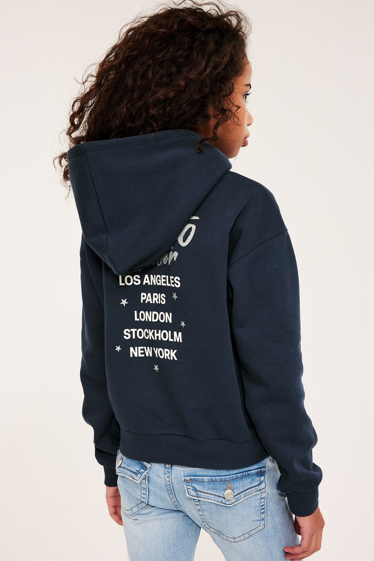 Widok z tyłu: dziecko w granatowej bluzie z kapturem z nadrukiem i jasnoniebieskich jeansach.