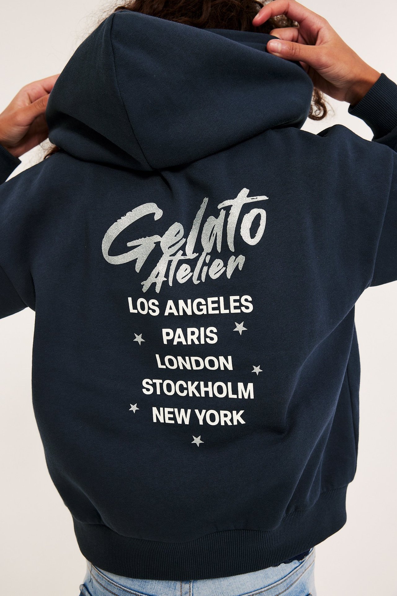 Widok z tyłu dziecka w granatowej bluzie z kapturem z srebrnym "Gelato Atelier" i białym nadrukiem miast.