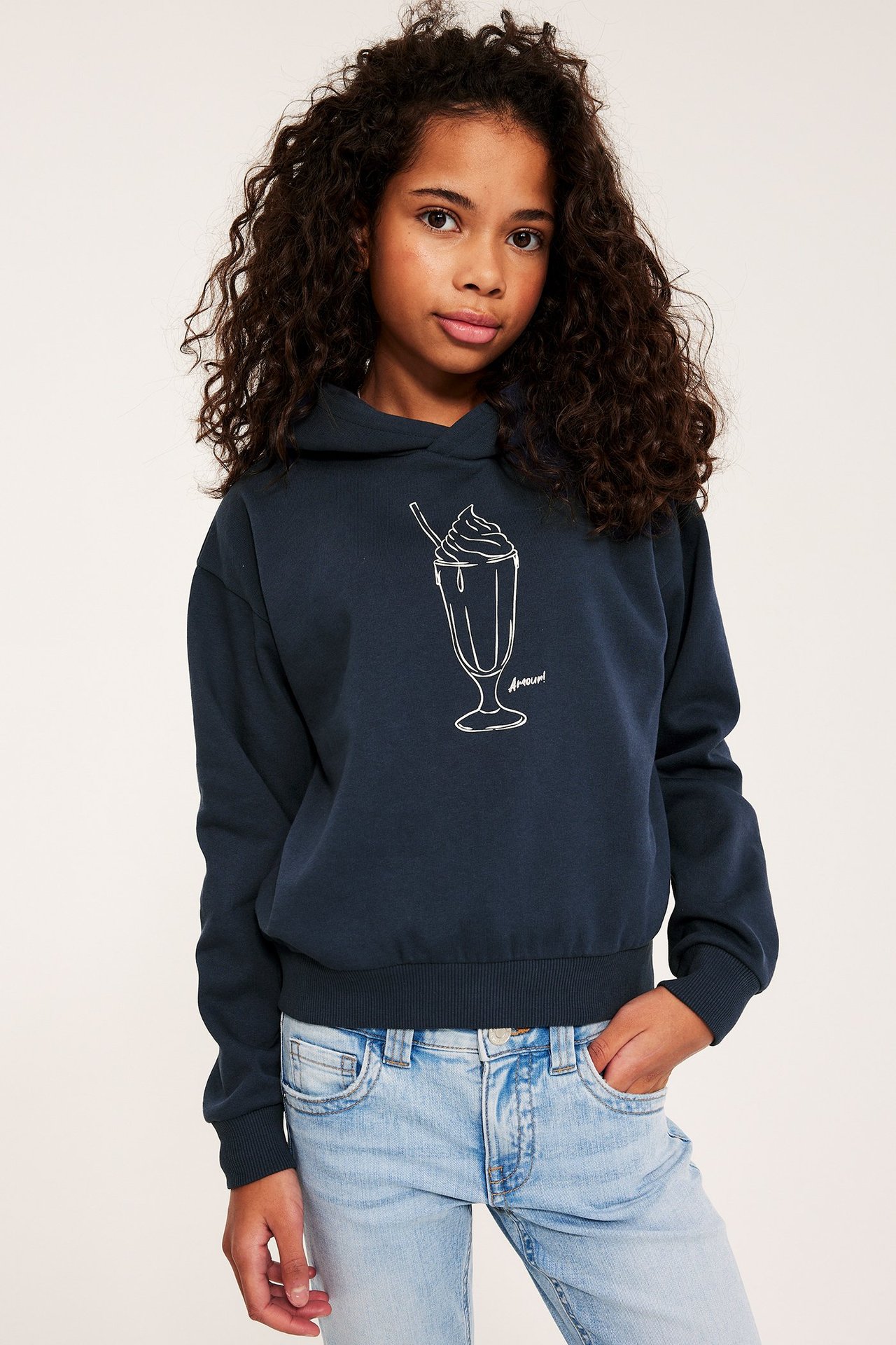 Frontbilde av et barn i blå hettegenser med hvitt milkshake-trykk og lyseblå jeans.