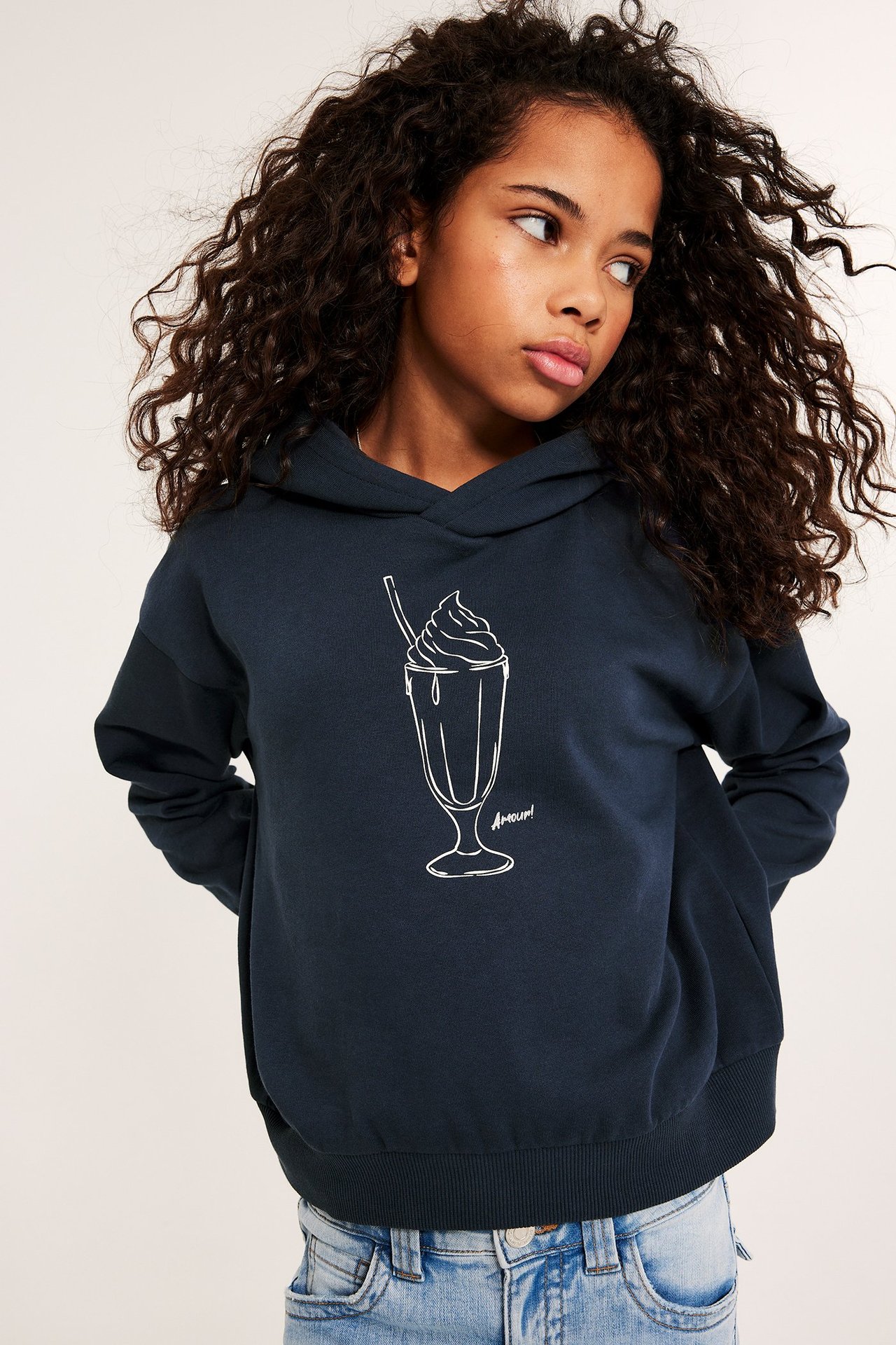 Frontvisning av et barn i mørkeblå hettegenser med hvitt milkshake-trykk, kombinert med lyseblå jeans.