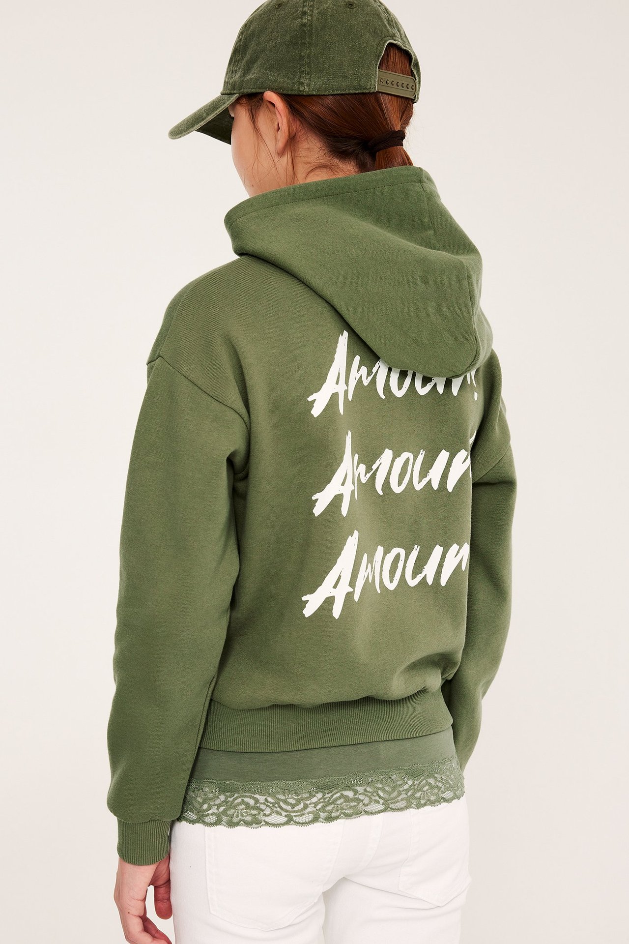 Dziecko tyłem: ciemnozielona bluza 'Amour', zielona czapka, białe spodnie, zielony koronkowy top.