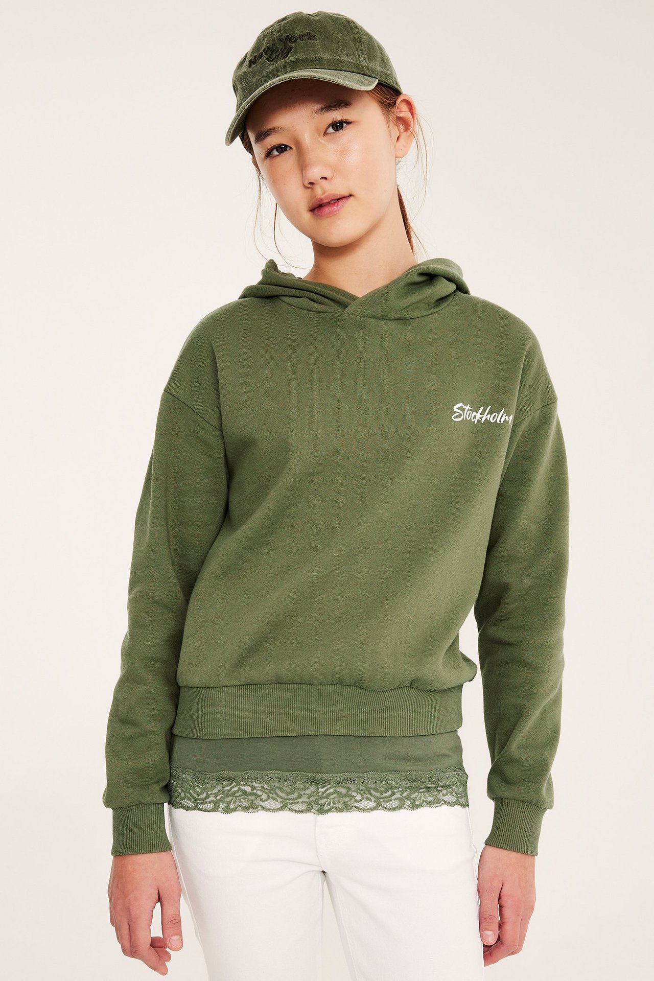 Edestä: Lapsi tummanvihreässä 'Stockholm'-hupparissa, vihreässä pitsitopissa, valkoisissa housuissa ja vihreässä lippiksessä.