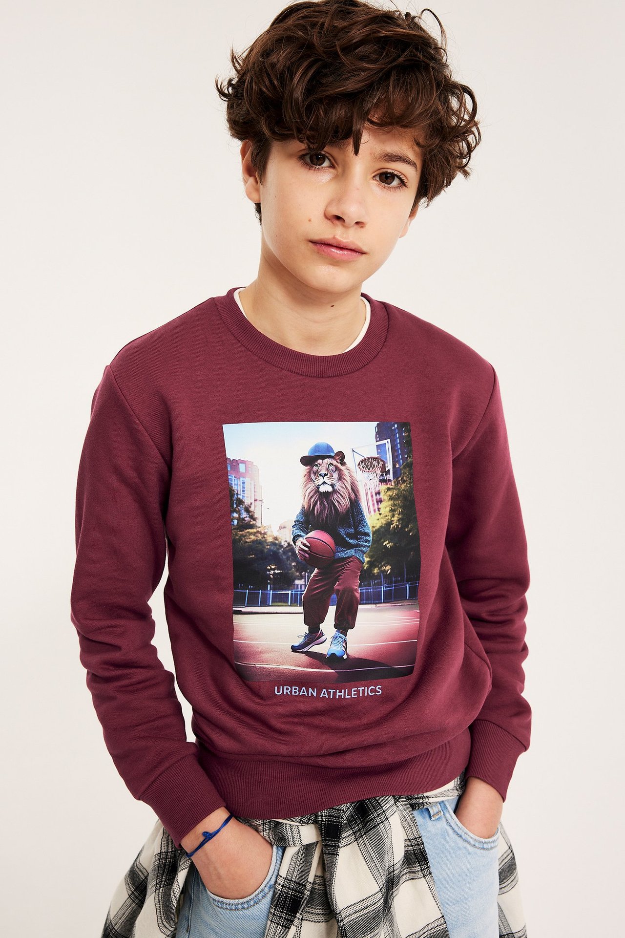 Framsida: Ett barn i vinröd sweatshirt med lejonbaskettryck, ljusa jeans och en rutig skjorta.