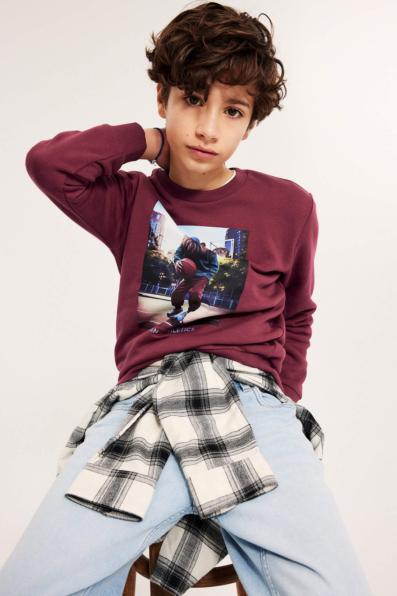 Widok z przodu, chłopiec w bordowej bluzie z nadrukiem koszykarza, jasnoniebieskie dżinsy, kraciasta koszula.