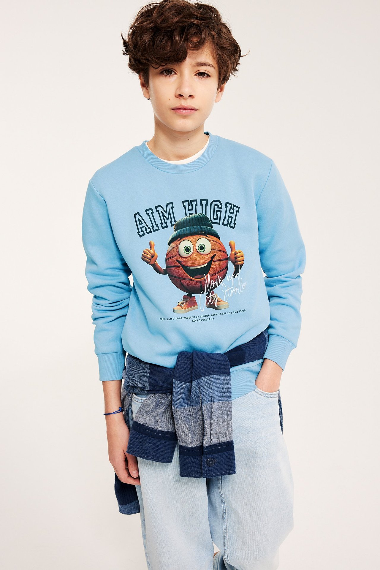 Kid bär ljusblå bomullssweatshirt med basketbolltryck, ljusa jeans och randig tröja knuten i midjan.