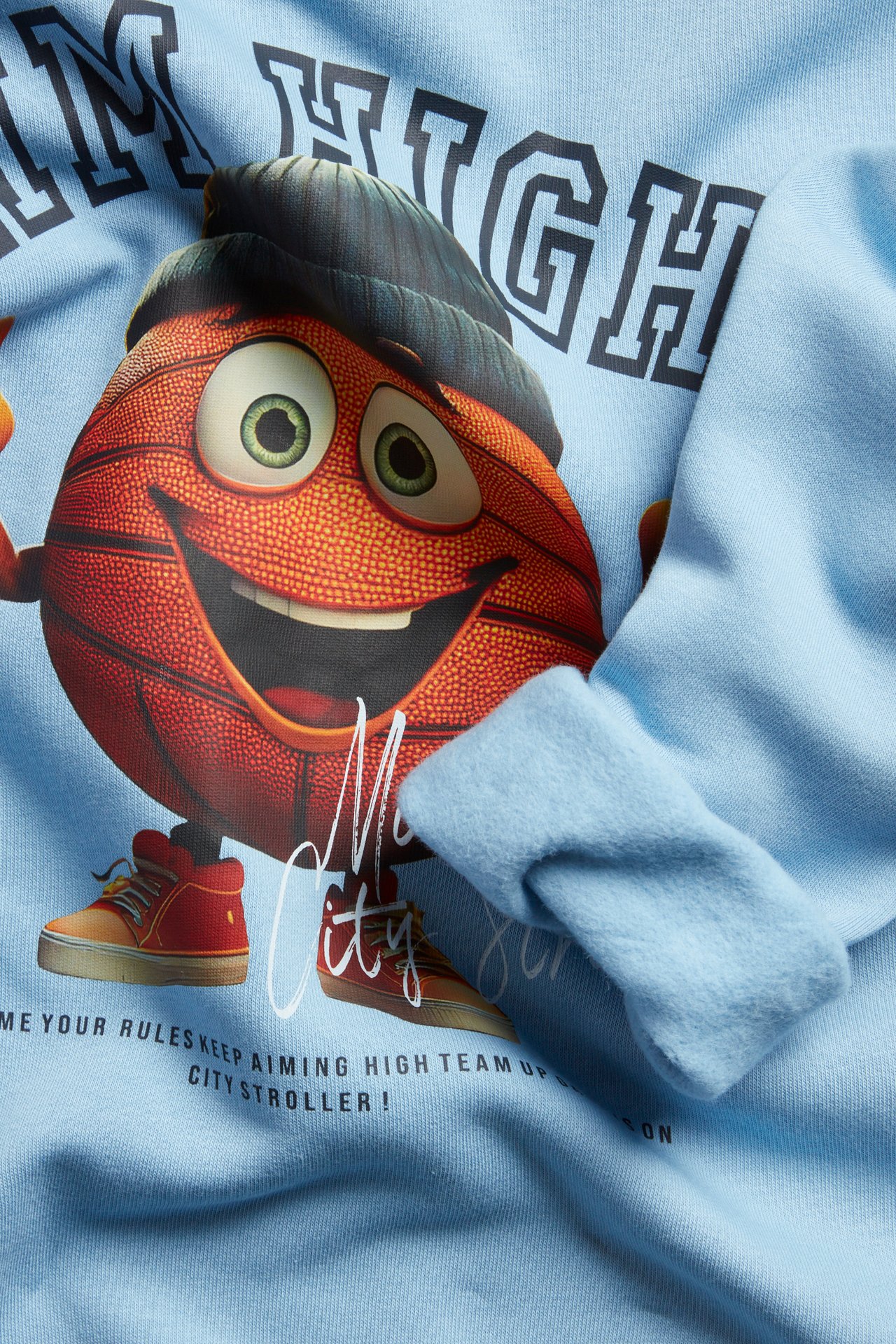 Zbliżenie na błękitną dziecięcą bluzę bawełnianą z nadrukiem uśmiechniętej postaci koszykówki i tekstem.