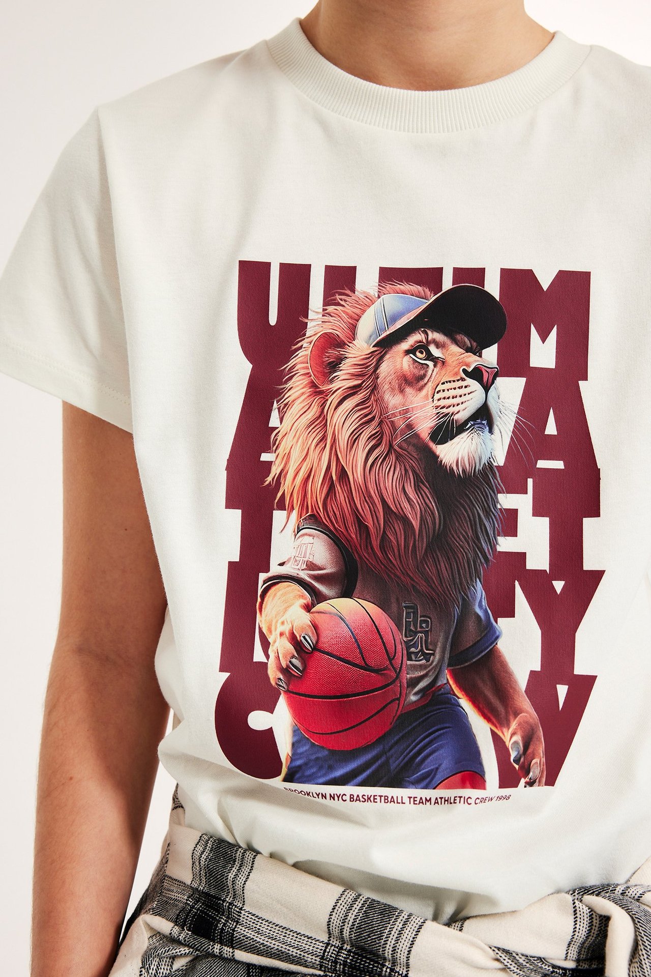 Lähikuva lapsen luonnonvalkoisesta puuvilla-t-paidasta, jossa koripalloa pelaava leijona-kuvio, stailattu ruutukauluspaidalla.