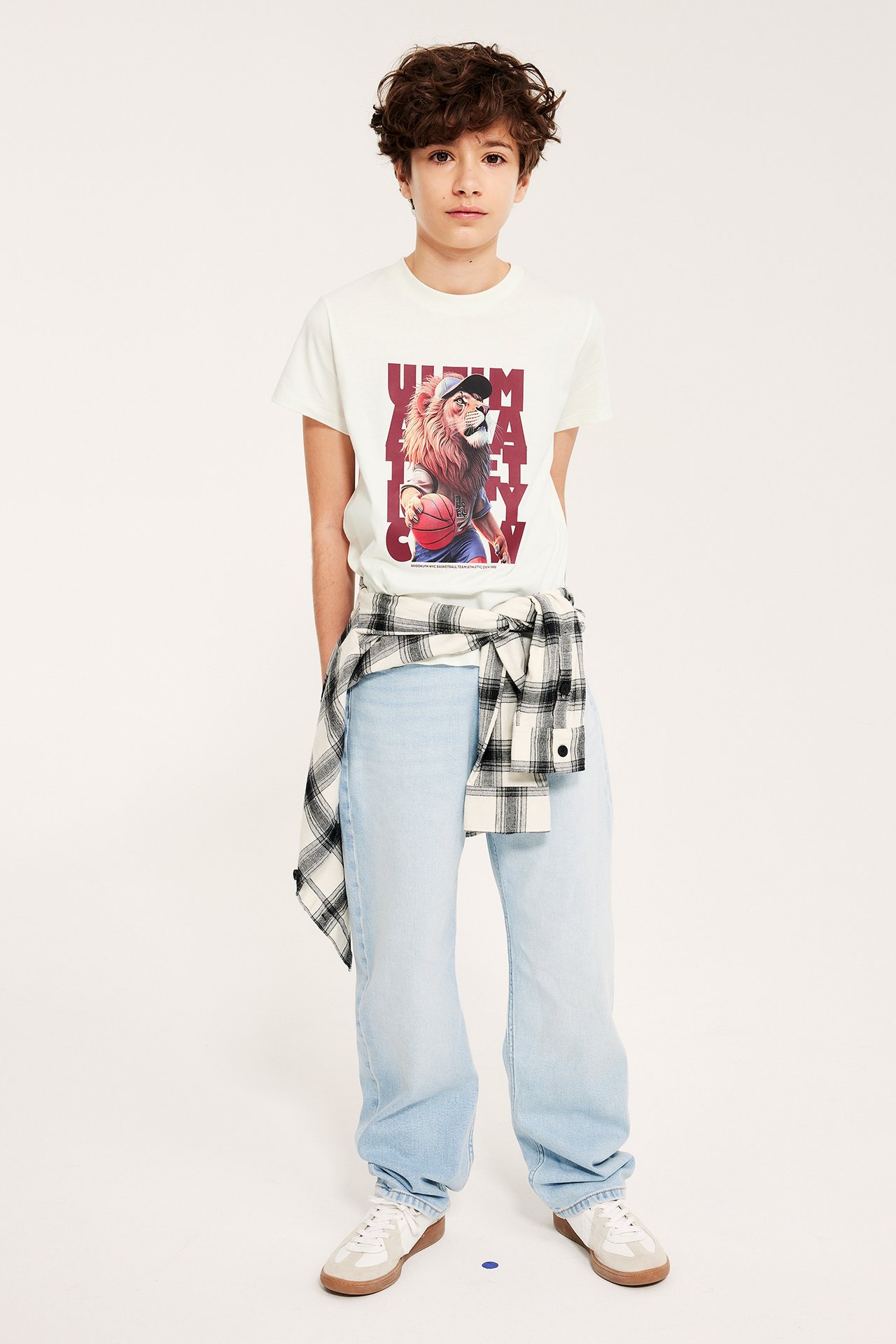 Frontutsikt av et barn i offwhite T-skjorte med løvetrykk, lyseblå jeans, og rutete skjorte knytt rundt midjen.