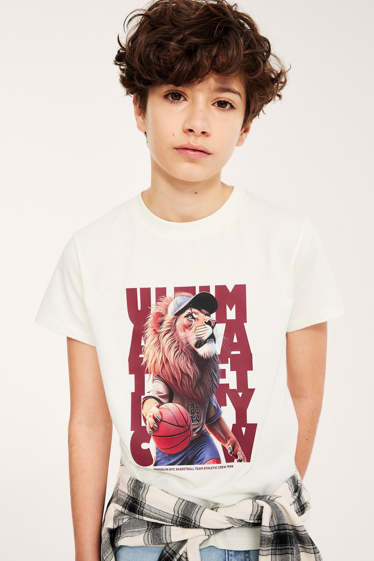 Dziecko w kremowym t-shircie z lwem-koszykarzem, koszula w kratę zawiązana w pasie.