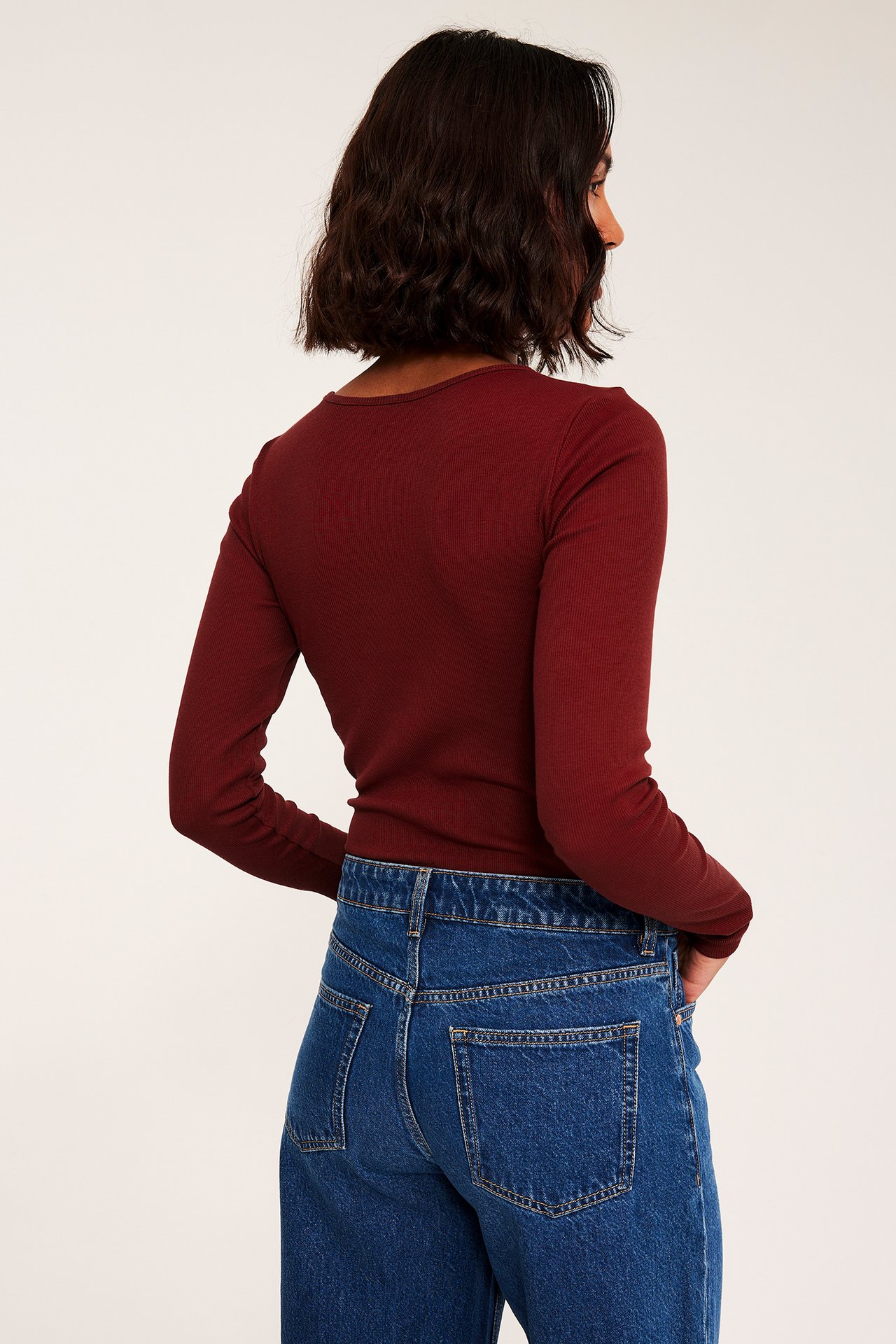 Kvinne sett bakfra/siden i en mørkerød ribbet langermet topp og blå denimjeans.