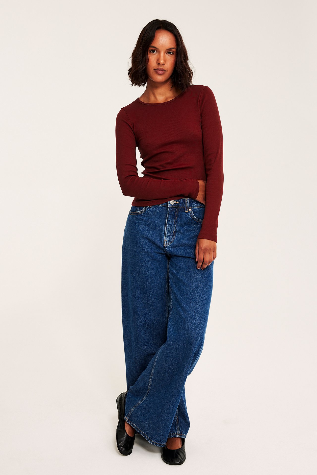 Frontvisning av kvinne i rød langermet topp, blå vide jeans og sorte ballerinasko.