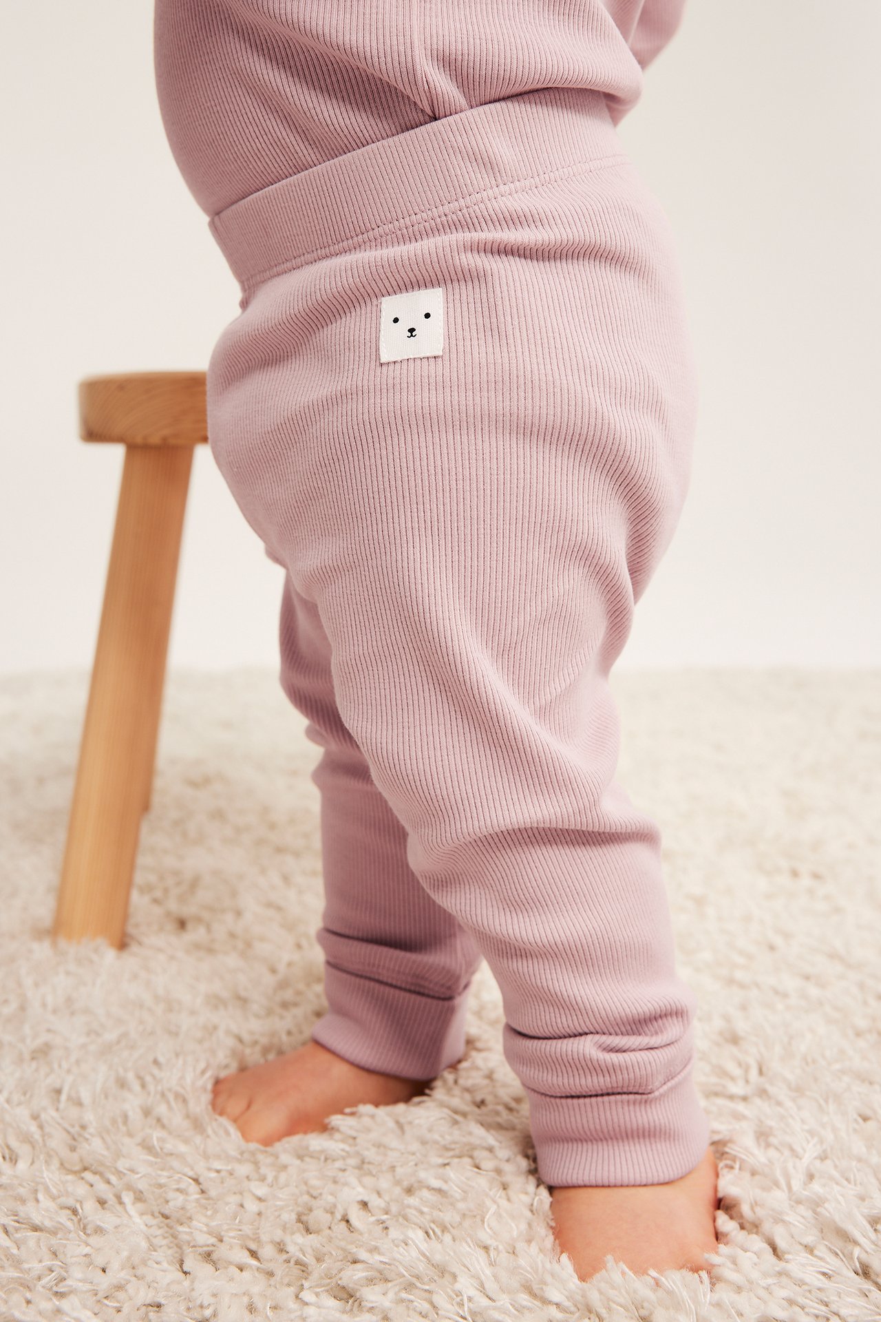 Sideprofil av baby i lilla ribbestrikkede leggings med hvitt bjørnemerke, stående barfot på et hvitt teppe.