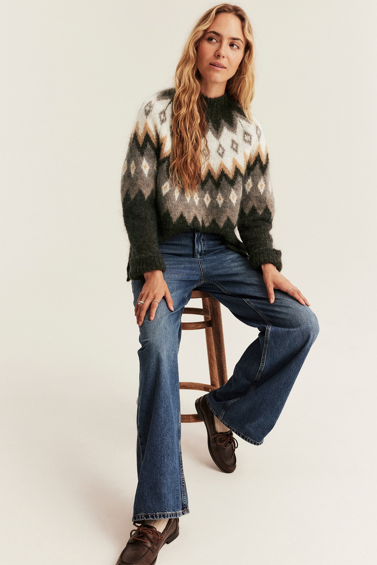 Frontbilde: kvinne i mørkegrønn, hvit, beige fair isle-genser, blå vide jeans, brune båtsko.