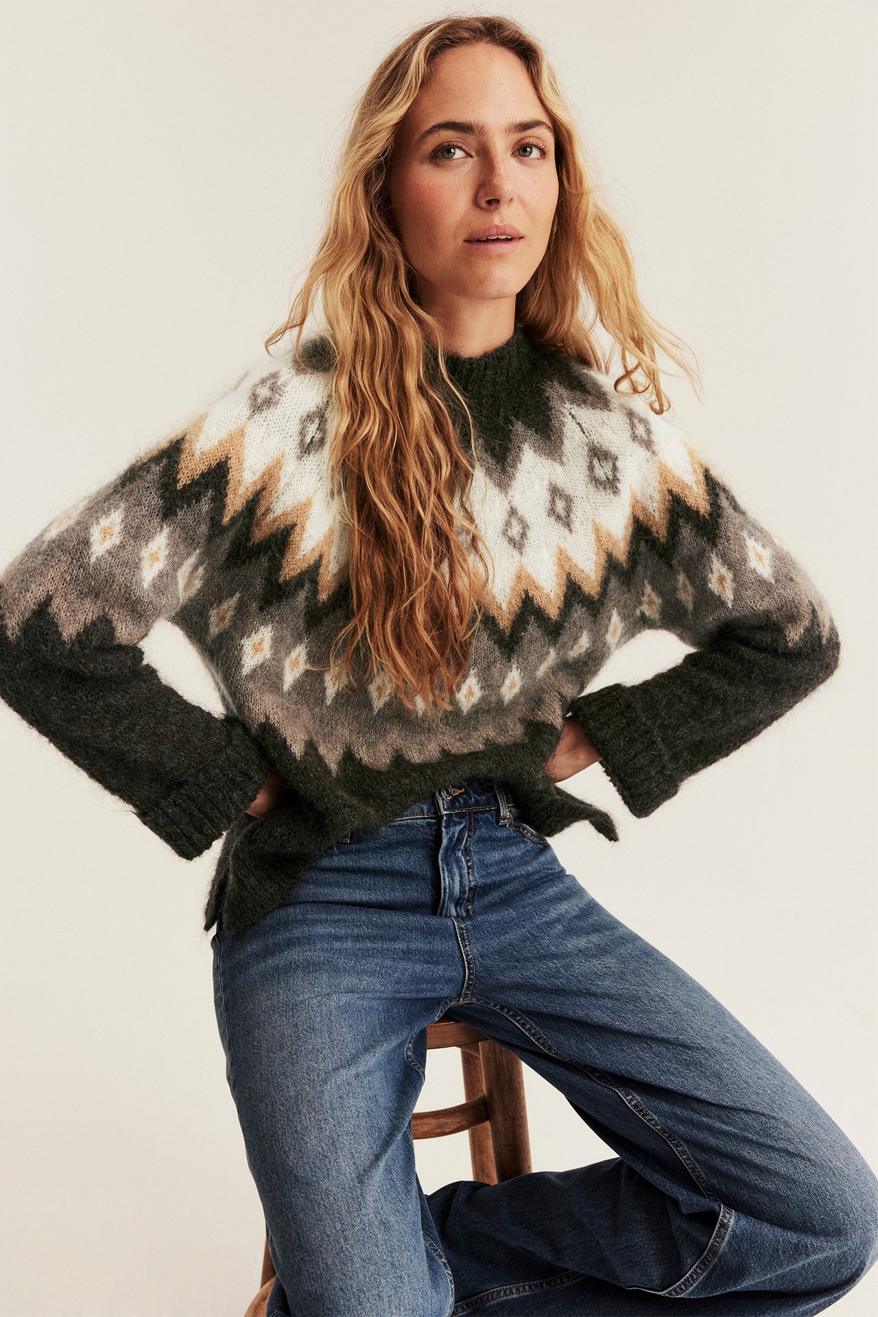 Kvinnelig modell i fair isle-strikkegenser (grønn, hvit, beige, grå) og blå jeans.