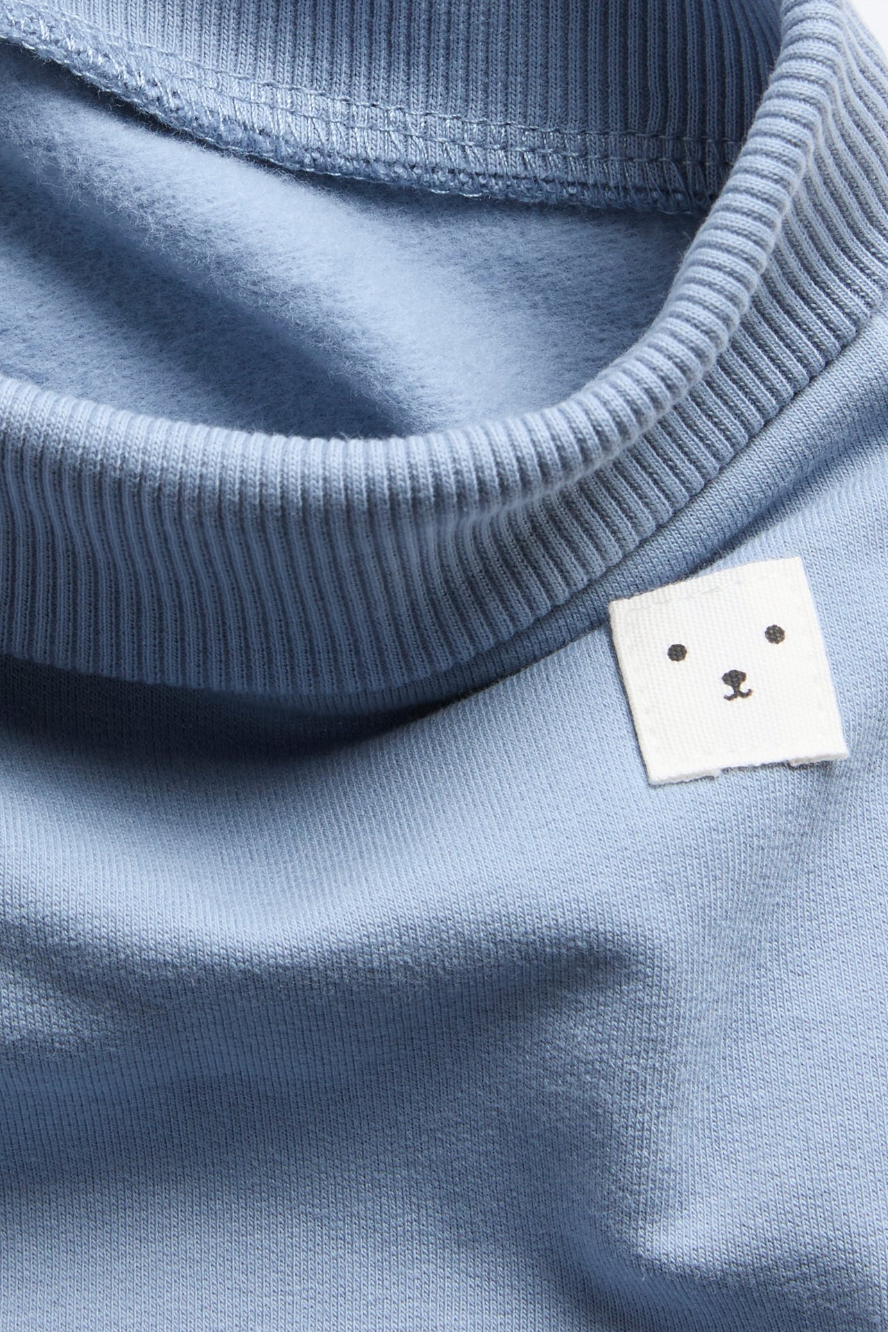 Lähikuva vaaleansinisestä lasten collegepaidasta, jossa ribbineulottu pääntie ja valkoinen karhupää-merkki.