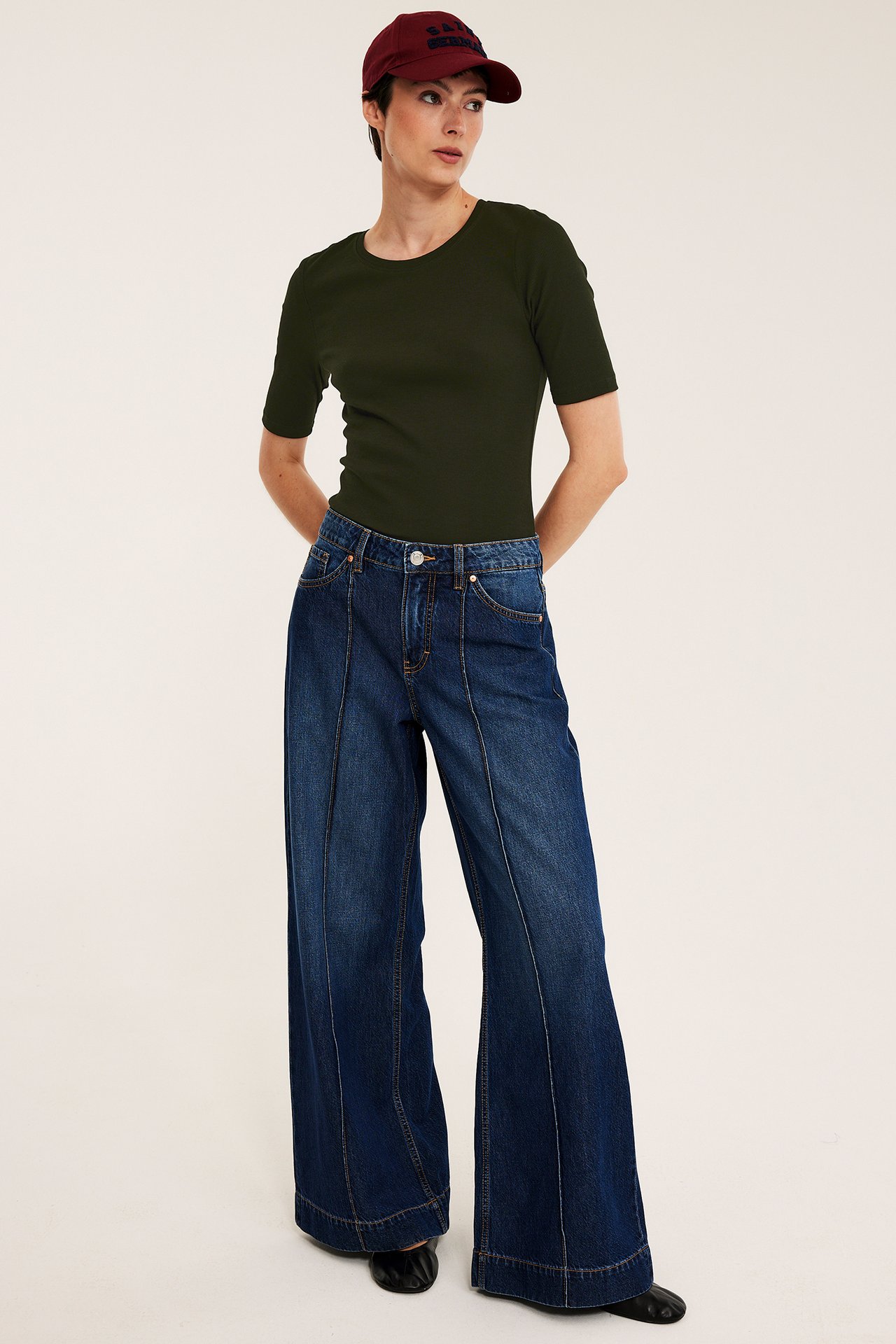 Framifrån, kvinna i grön ribbad kortärmad topp, blå vida jeans, vinröd keps och svarta ballerinaskor.