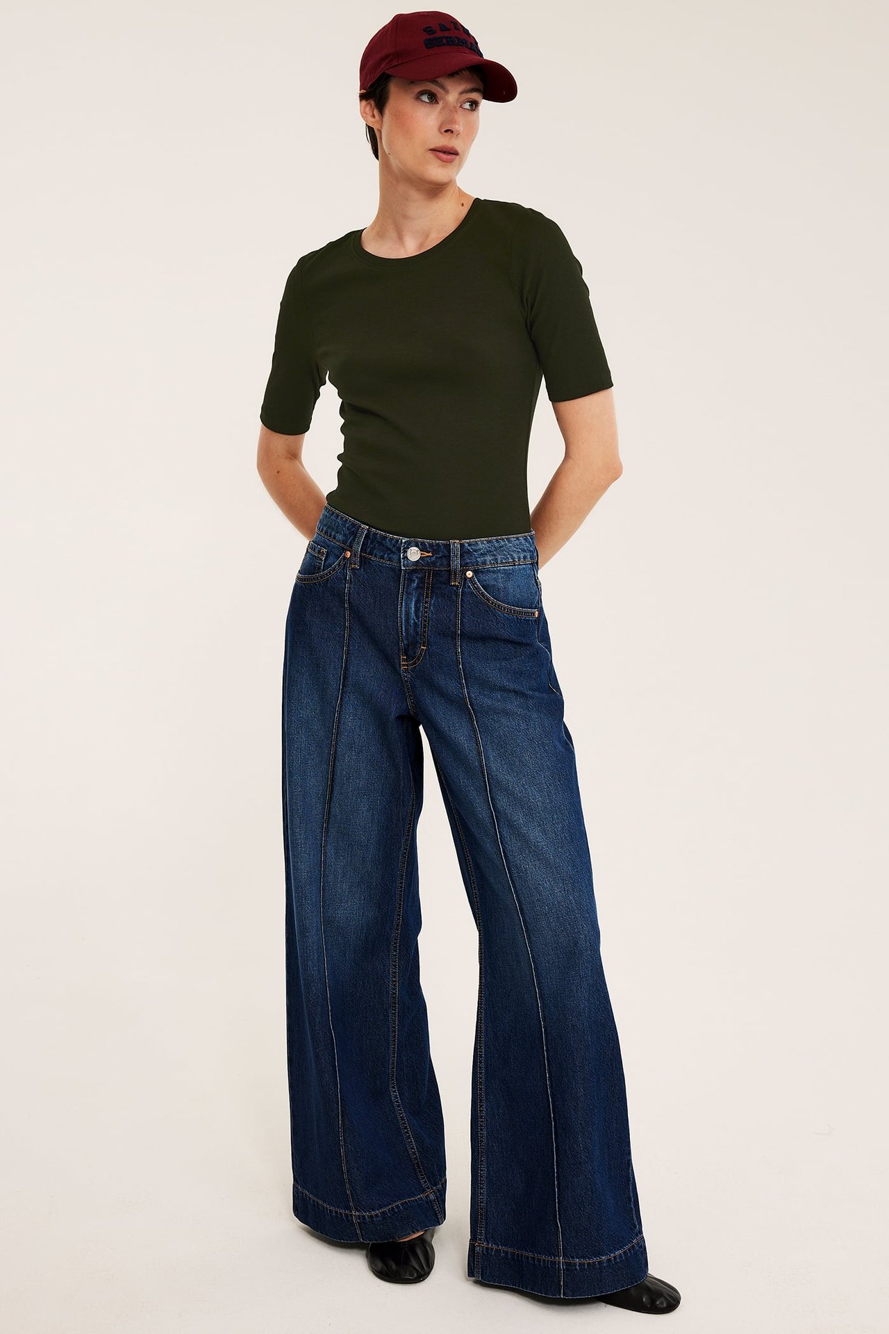 Framifrån, kvinna i grön ribbad kortärmad topp, blå vida jeans, vinröd keps och svarta ballerinaskor.