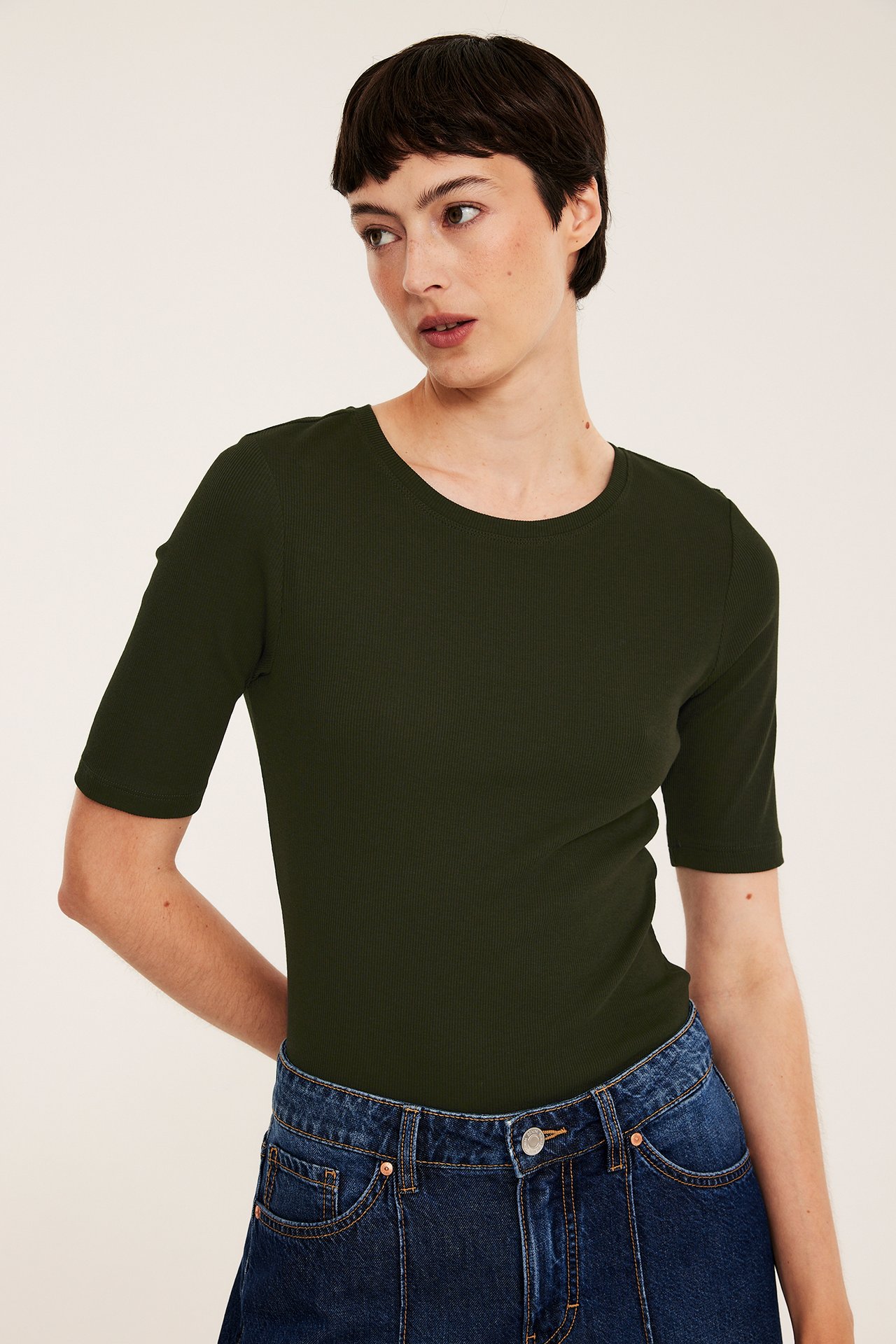 Framifrån, kvinna i mörkgrön ribbad kortärmad topp och mörkblå jeans.
