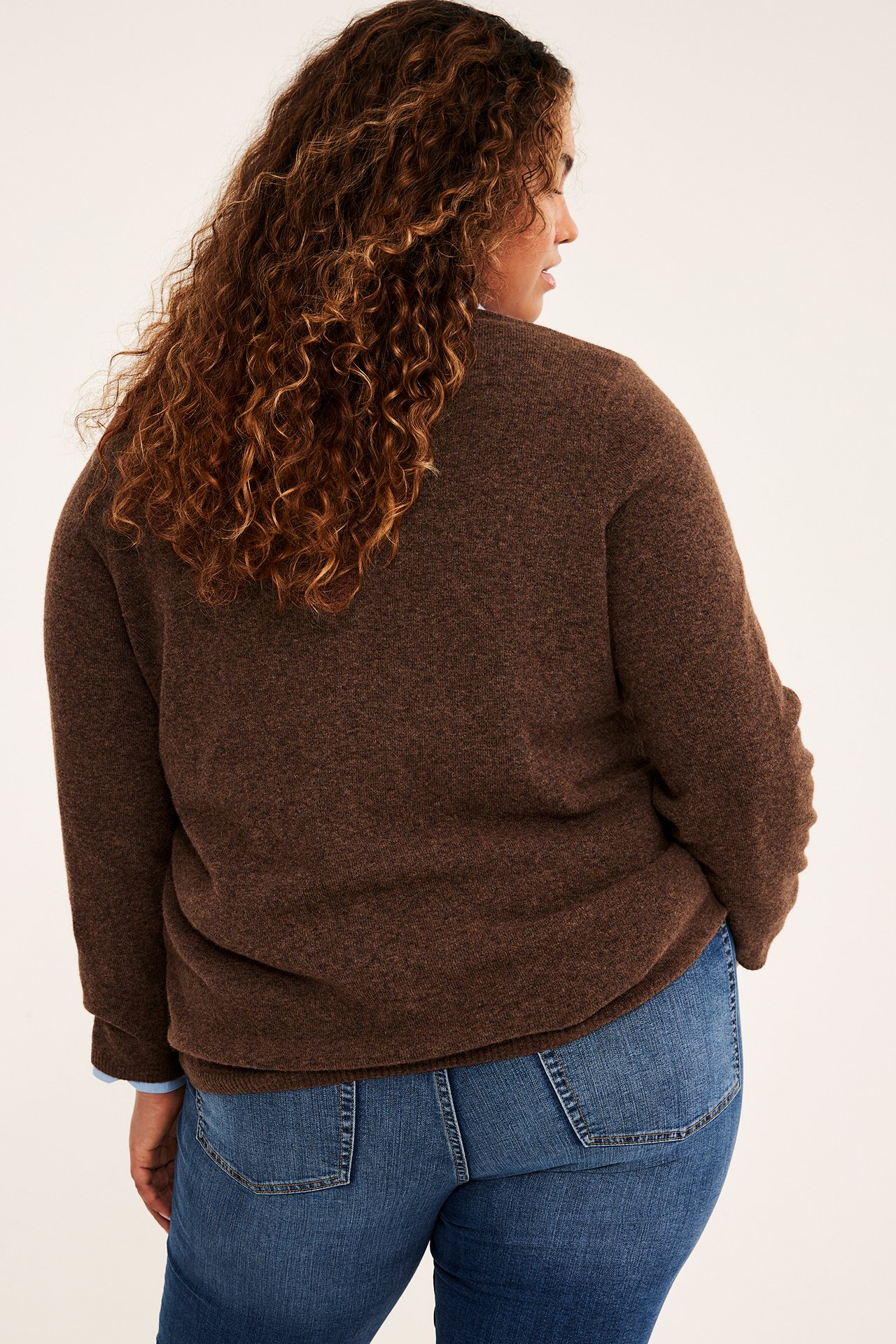 Kvinne sett bakfra i brun, teksturert, løs genser over blå skjorte og blå jeans.