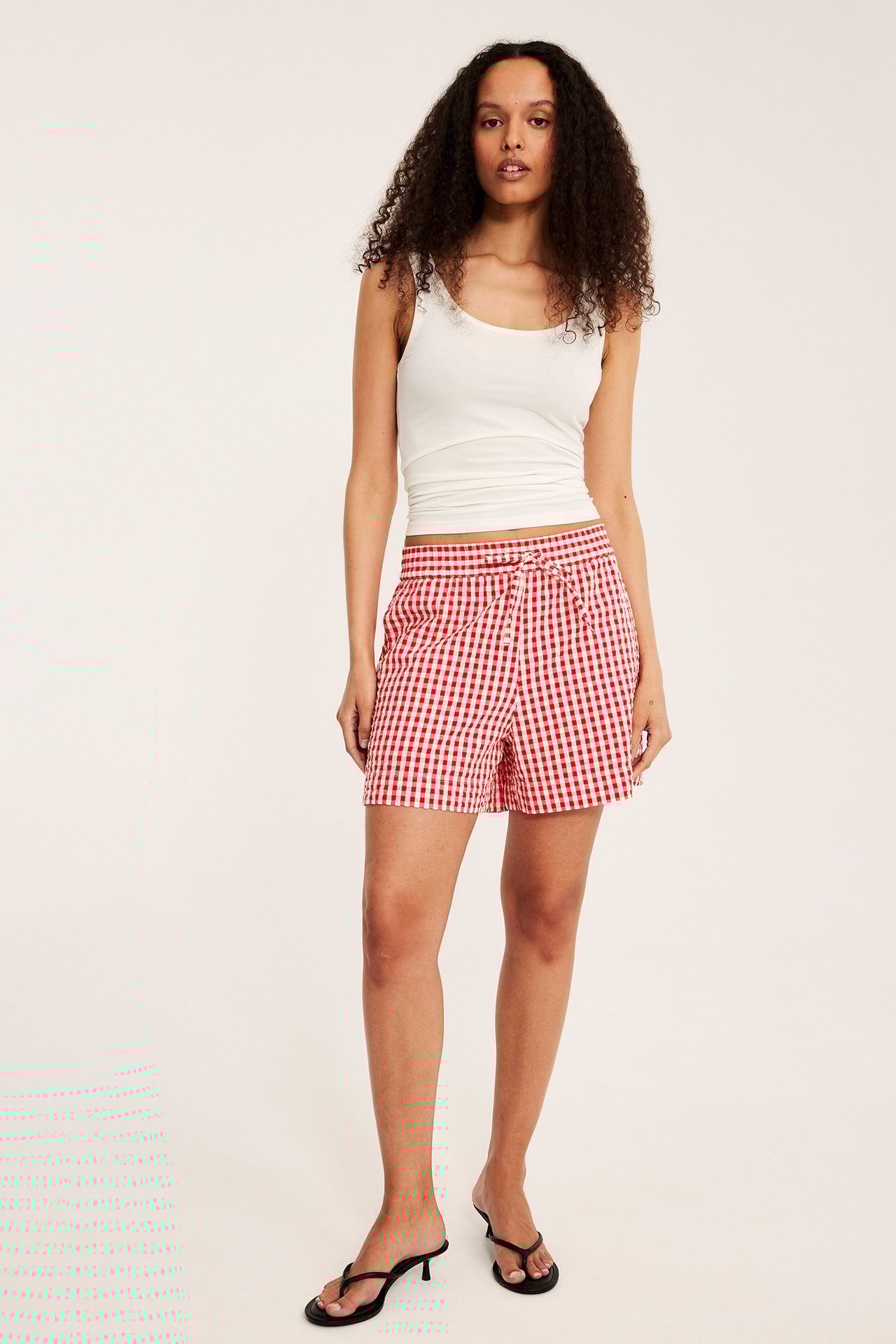 Widok z przodu: Kobieta w czerwonych kratkowych szortach, białym topie i czarnych japonkach.
