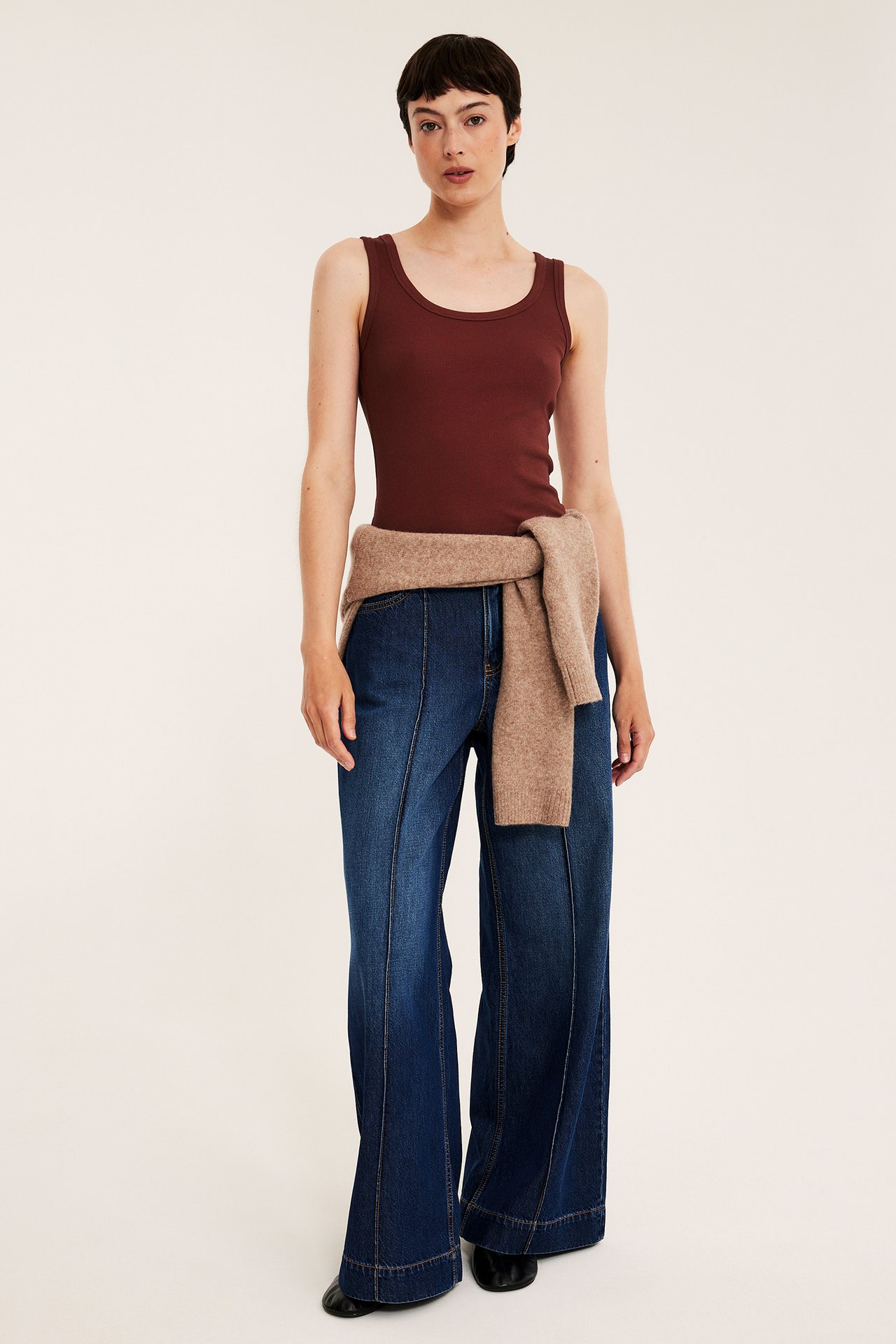 Framsida, kvinna i mörk röd ribbad tanktopp, mörkblå vida jeans och ljusbrun tröja.