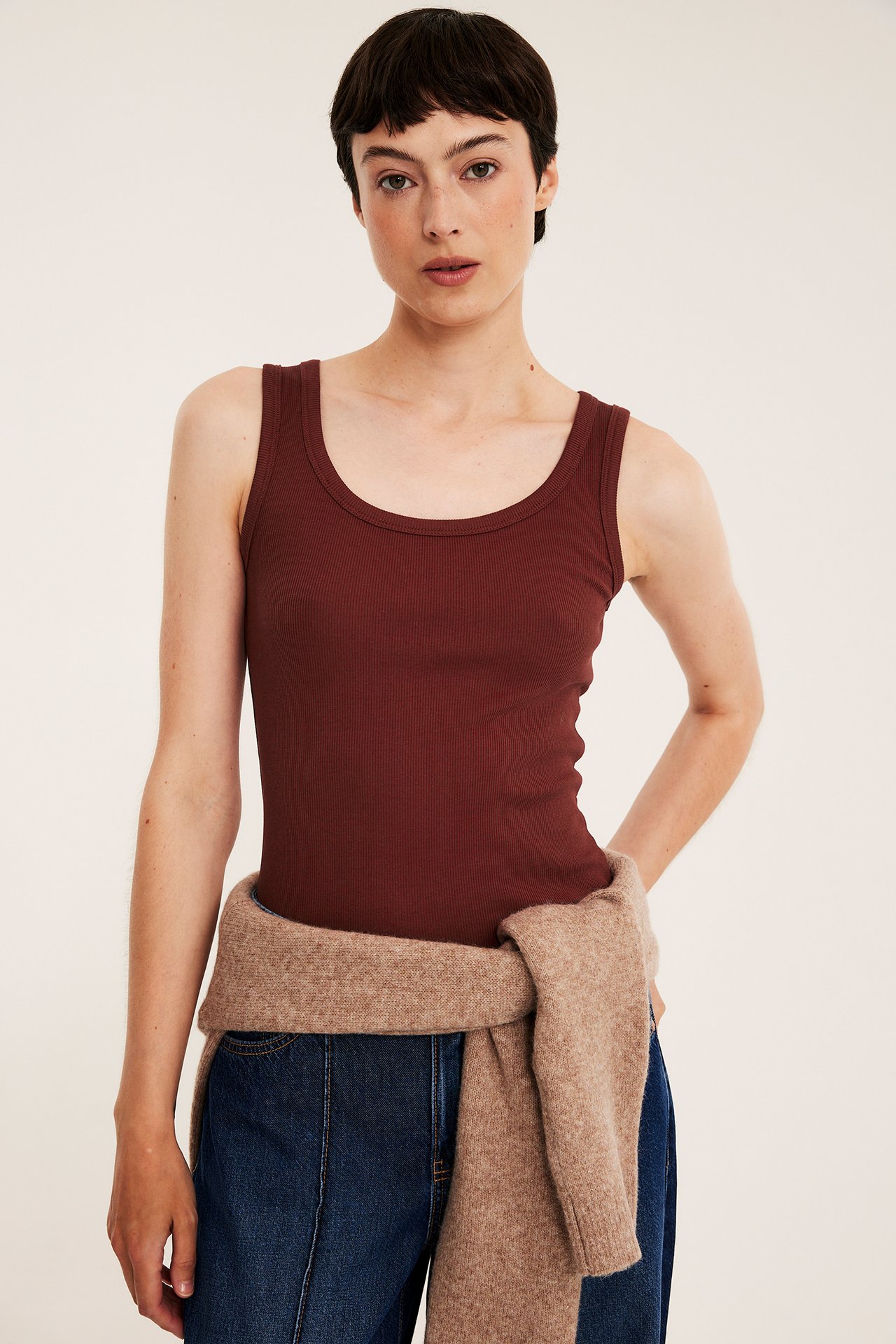 Frontvy av kvinna i mörkröd ribbad tanktopp, blå jeans och beige tröja knuten i midjan.