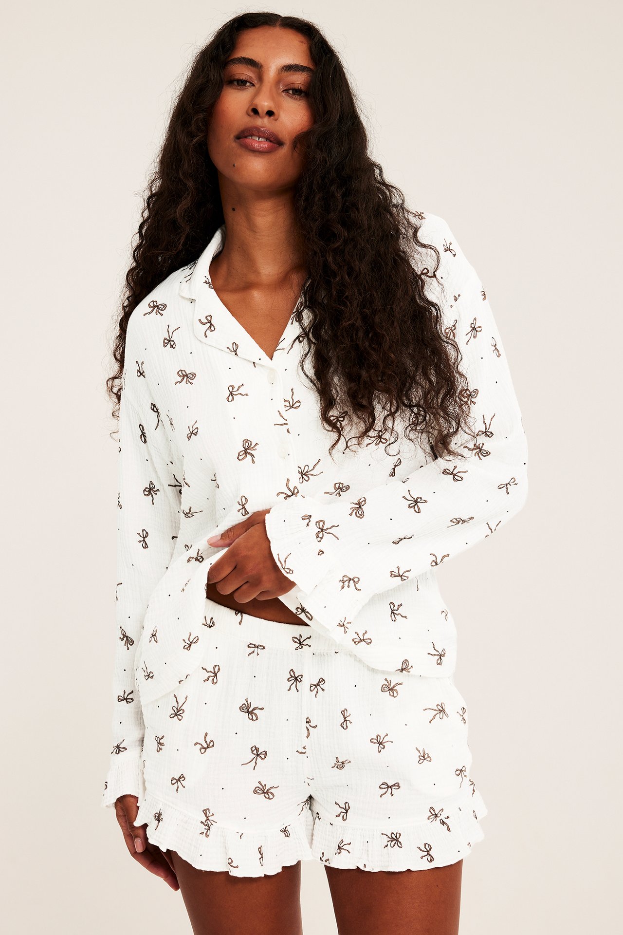 Edestä: Naishenkilö valkoisessa pyjamasetissä ruskealla rusettiprintillä ja röyhelöshortseilla.