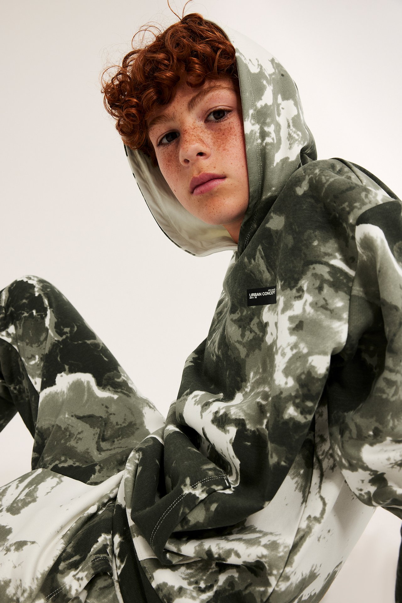 Kid bærer en løs tie-dye mørkegrønn og hvit hettegenser og matchende joggebukser, forfra.