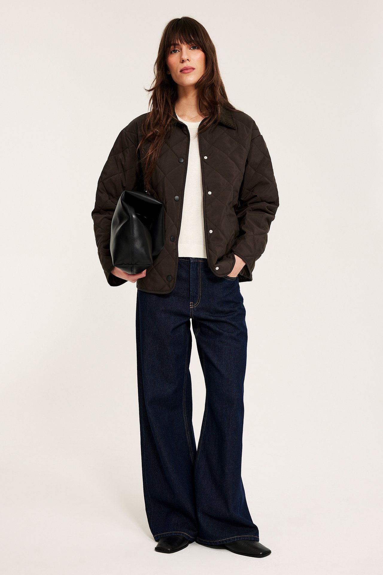 Kvinna i mörkbrun quiltad jacka med manchesterkrage, vit topp, blå jeans, håller en svart väska.