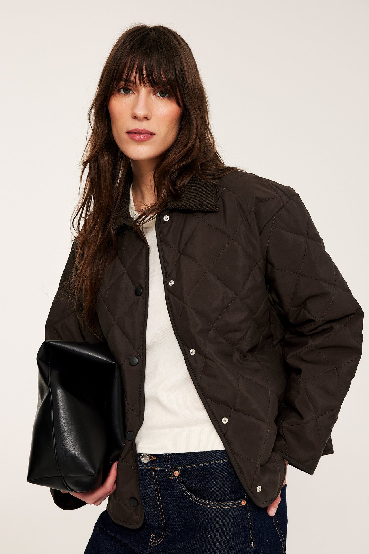 Nainen tummanruskeassa tikkitakissa, vakosamettikaulus, valkoinen neule, tummat farkut, musta käsilaukku.