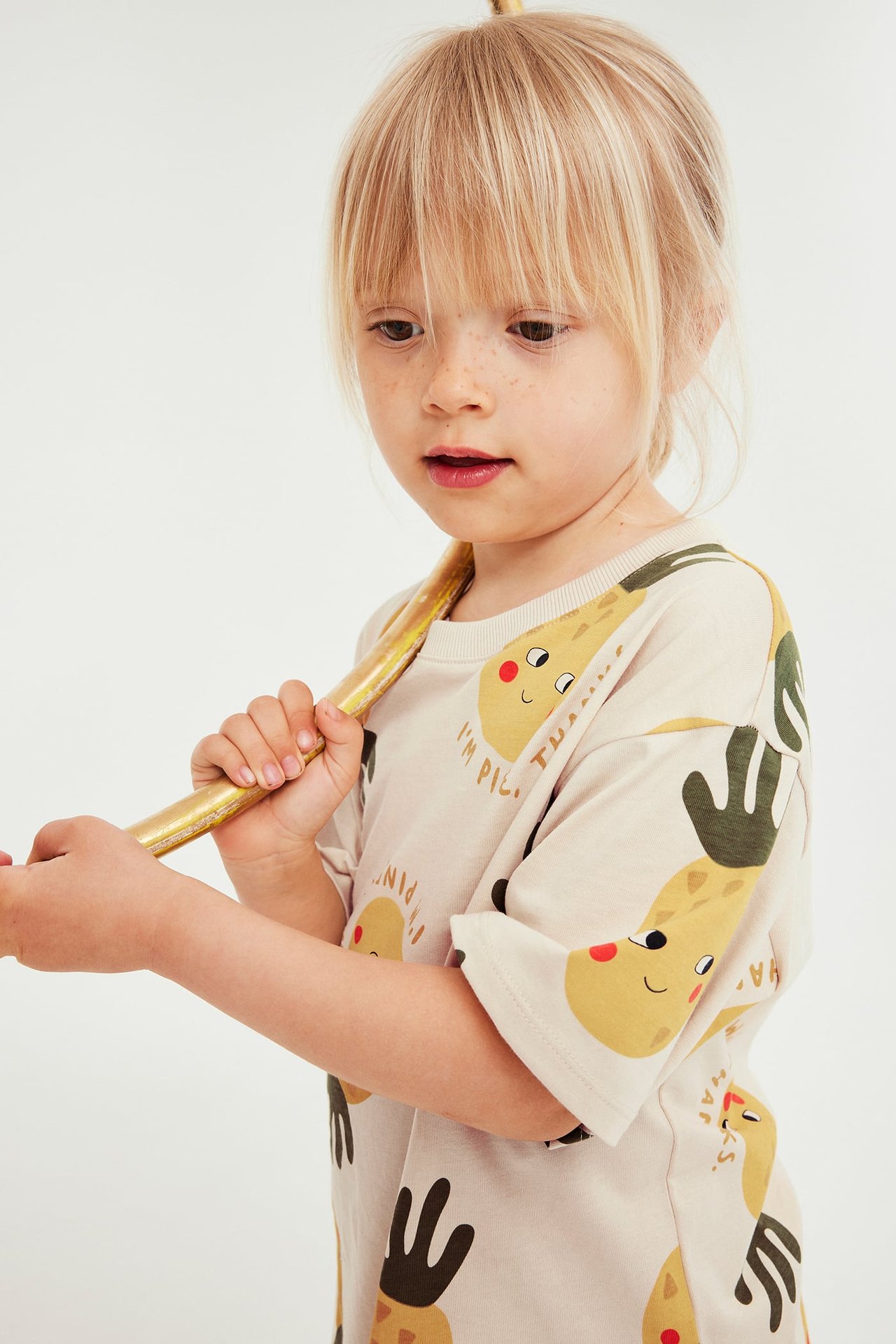 Forfra et barn i en lys beige t-skjorte med gult tegneserie-fruktmønster, holdende en gyllen pinne.