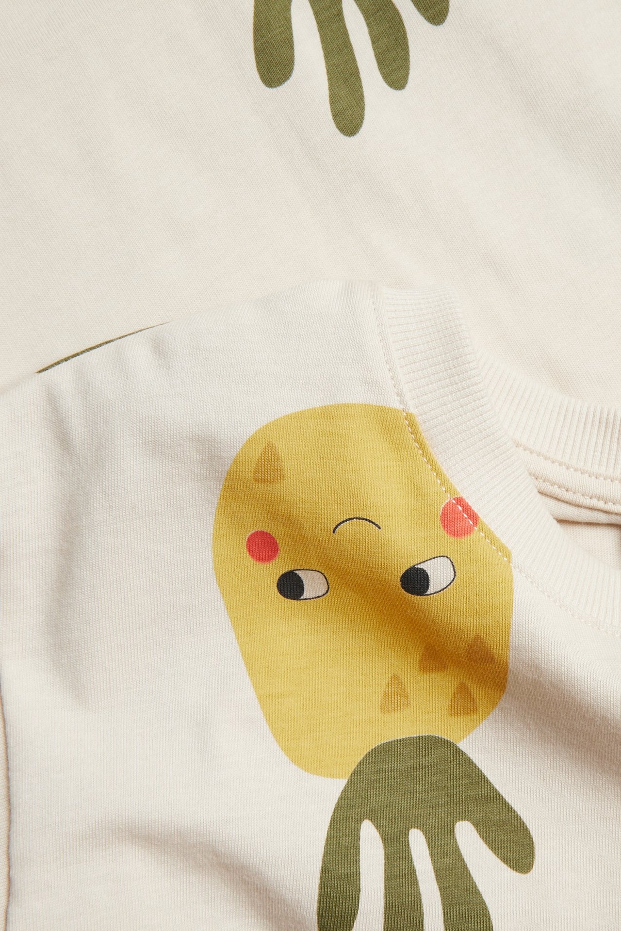Zbliżenie na bawełniany T-shirt dziecięcy w kolorze écru z żółtym nadrukiem owocowej postaci i oliwkowozielonymi kształtami.