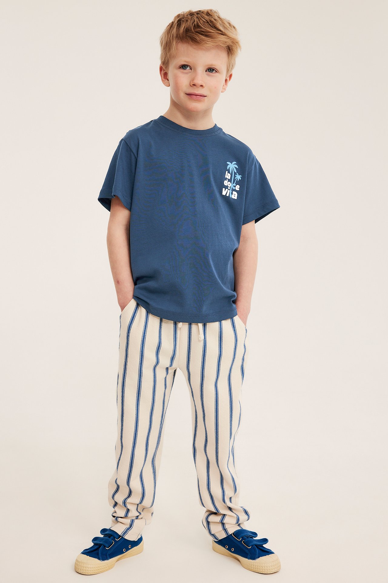Edestä: Lapsi sinisessä printti-t-paidassa, kermanväriset raidalliset housut ja siniset kengät.