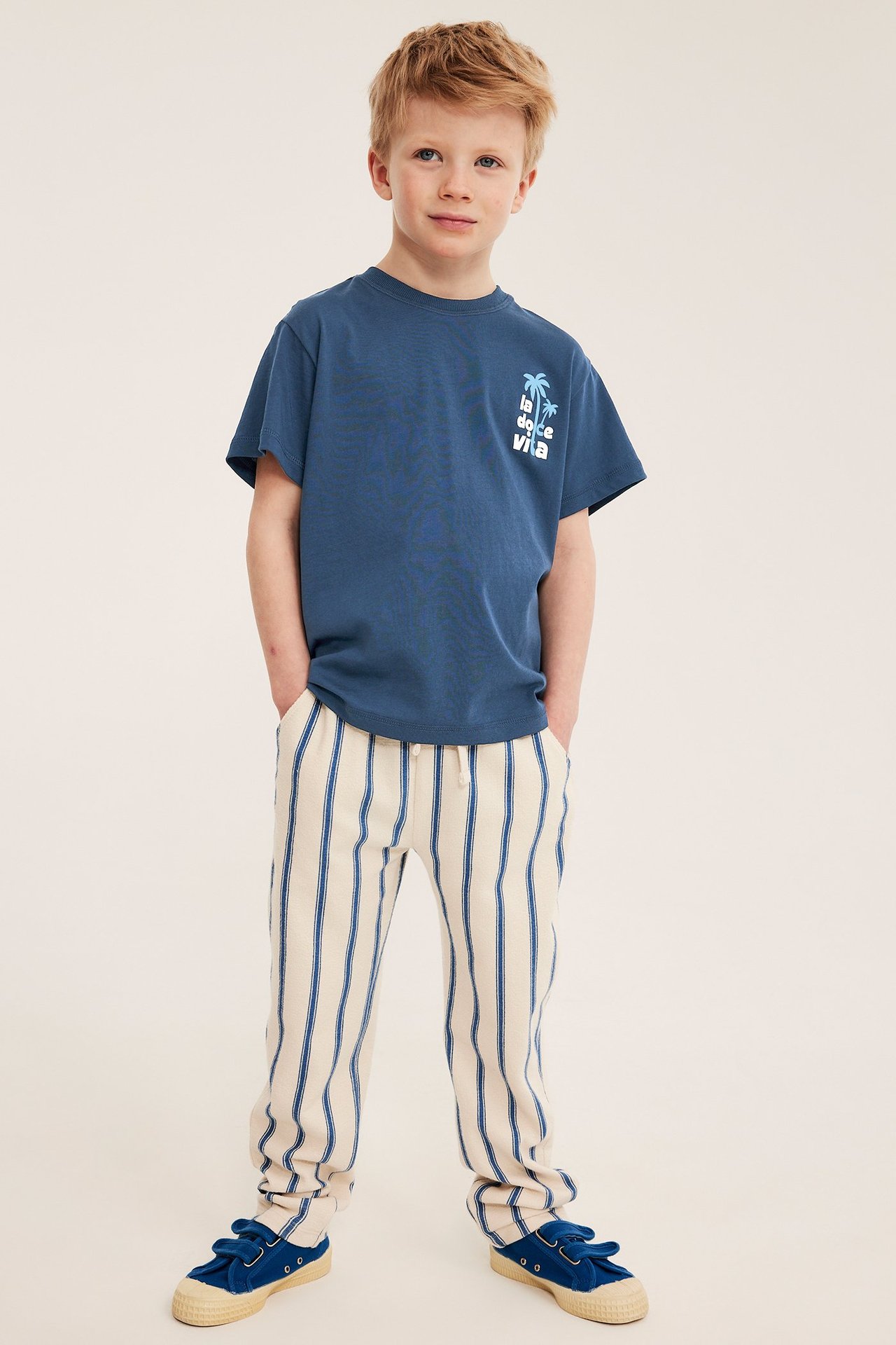 Edestä: Lapsi sinisessä printti-t-paidassa, kermanväriset raidalliset housut ja siniset kengät.