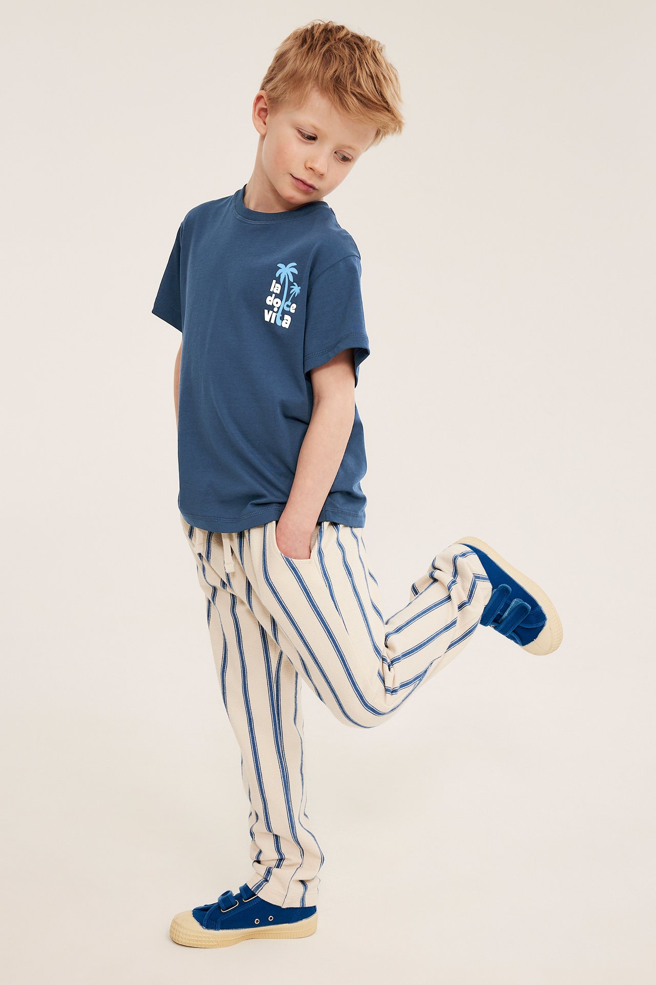Barn modellerer en blå oversized bomulls-t-skjorte med brysttrykk, kremfargede stripete bukser og blå canvassko.