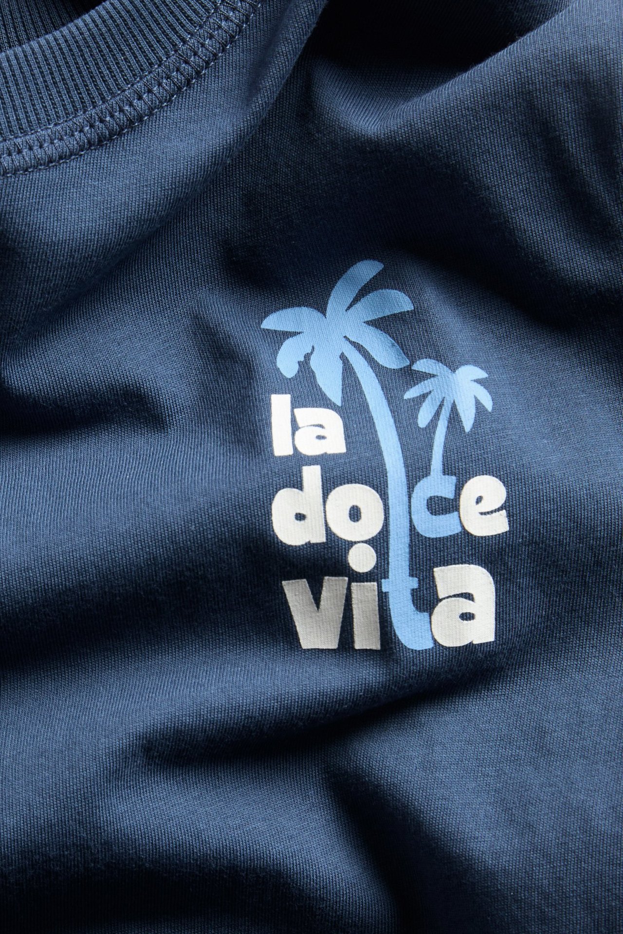 Zbliżenie na niebieską bawełnianą koszulkę z białym i jasnoniebieskim napisem 'La Dolce vita' oraz palmami.