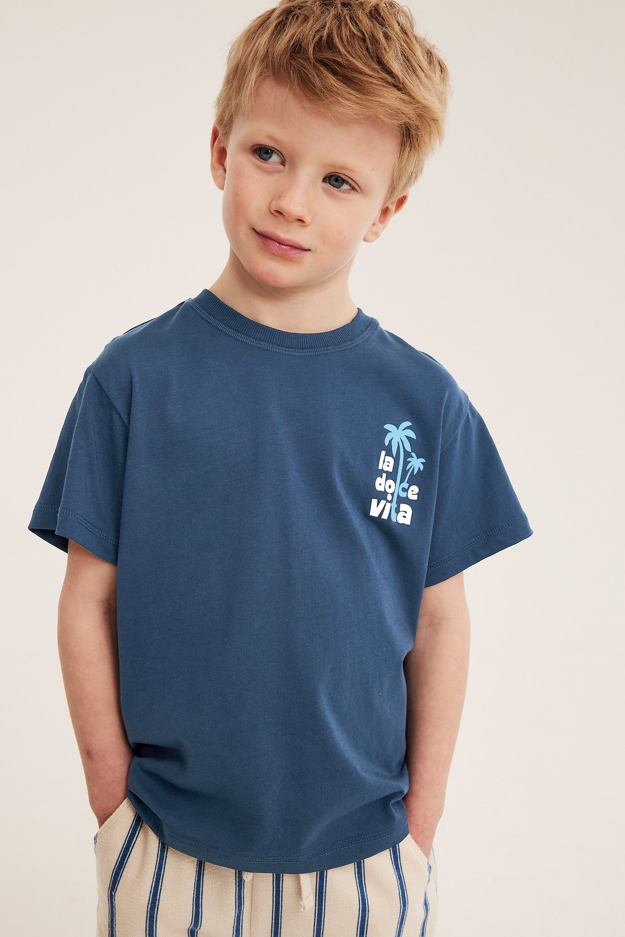 Widok z przodu: dziecko w niebieskim bawełnianym t-shircie oversize z nadrukiem "La Dolce Vita" i w paski spodniach.