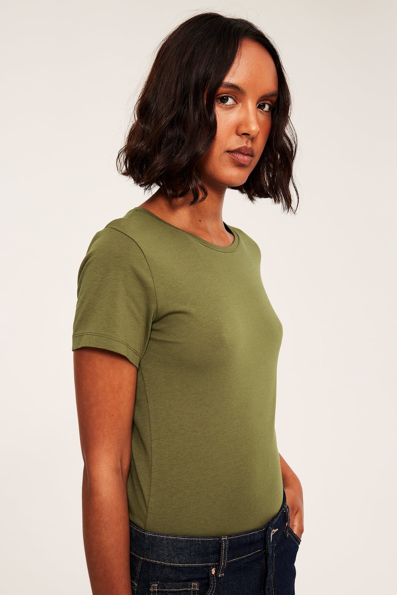 Sidovy av en kvinna som bär en åtsittande olivgrön kortärmad t-shirt och mörkblå jeans.