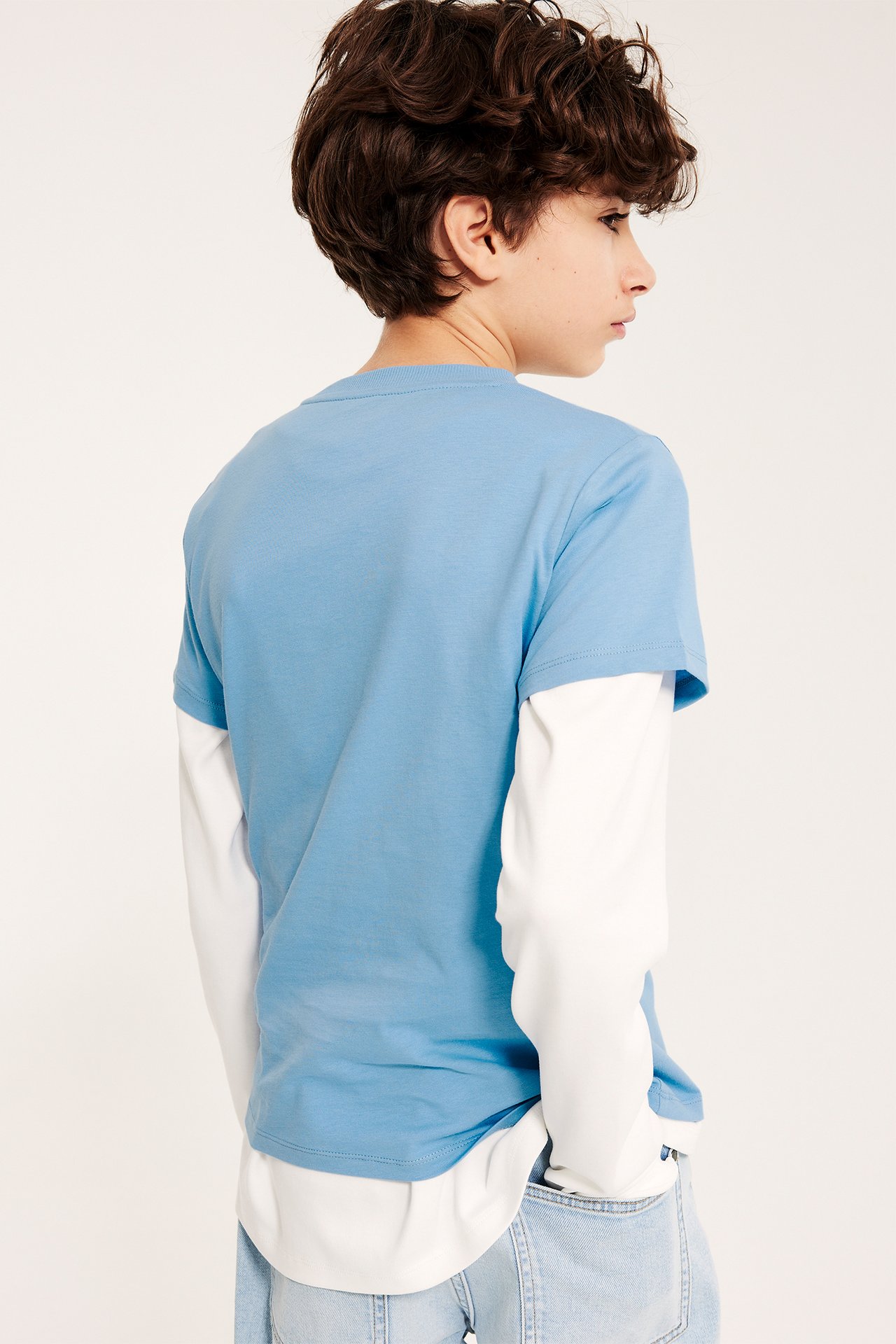 Sidebilde av et barn i lyseblå kortermet t-skjorte over hvit langermet genser og lyseblå jeans.
