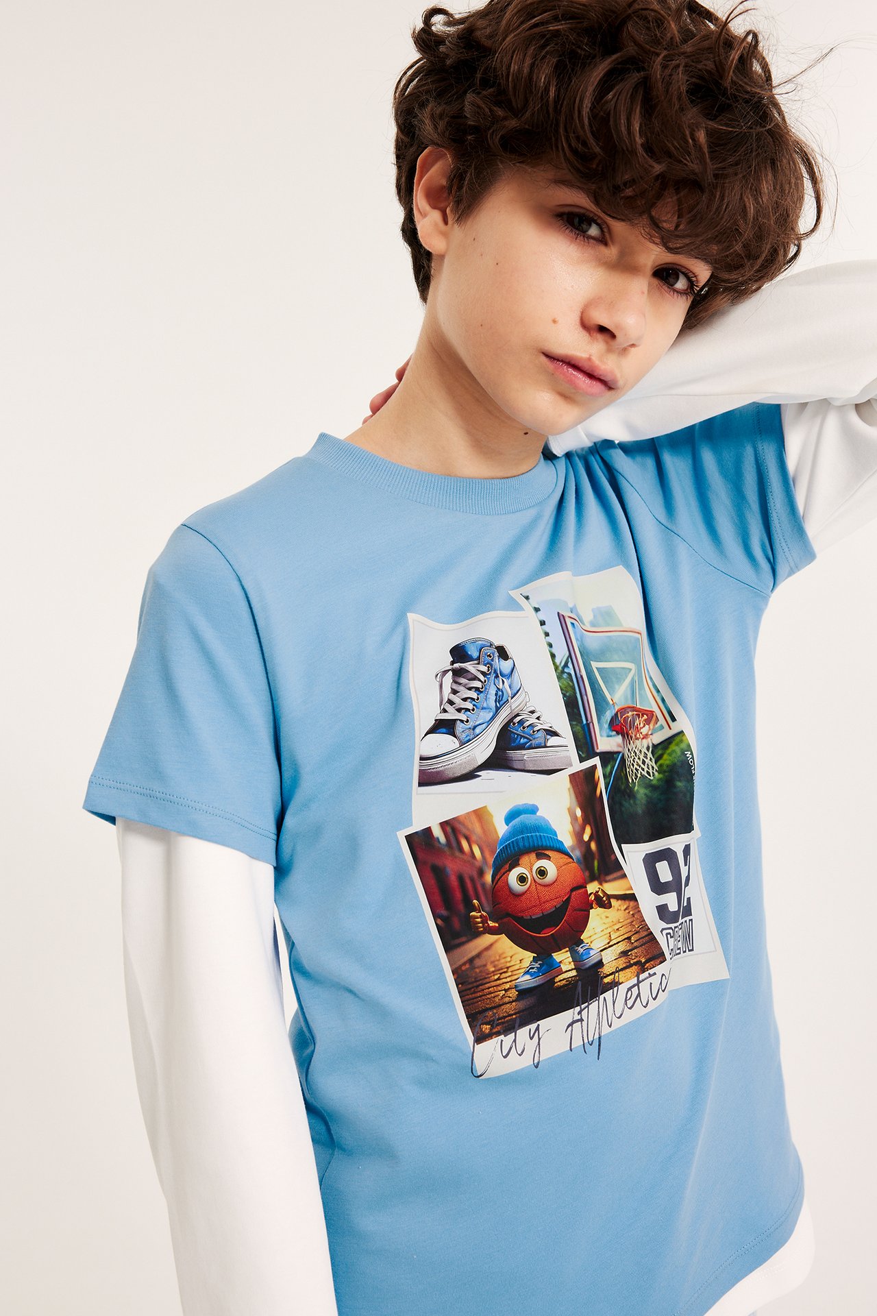 Edestä: Lapsi yllään vaaleansininen puuvilla t-paita koripallopainatuksella valkoisen pitkähihaisen päällä.