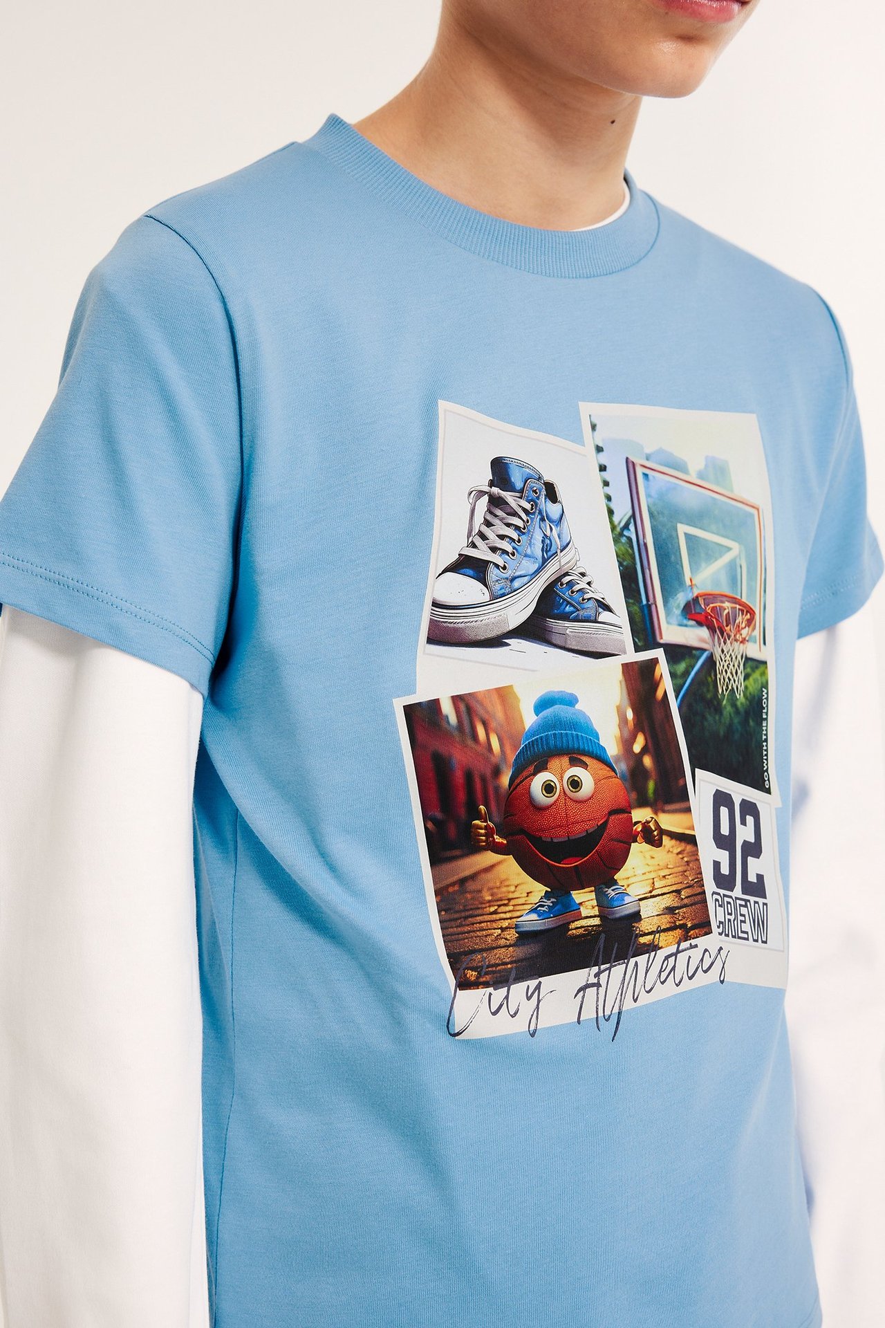 Frontvisning, barnas lyseblå bomulls-t-skjorte med basketballtrykk, over en hvit langermet trøye.