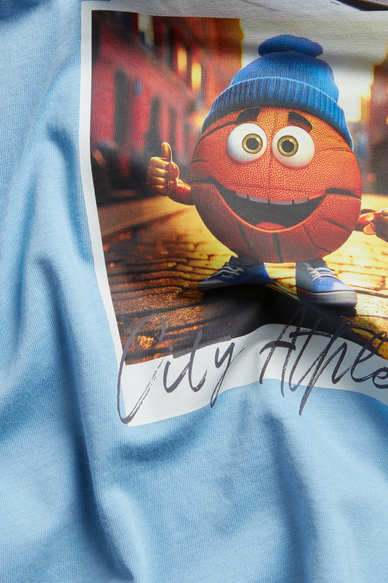Närbild: ljusblå bomulls-t-shirt för barn med stort tryck av en leende basketboll i blå mössa.