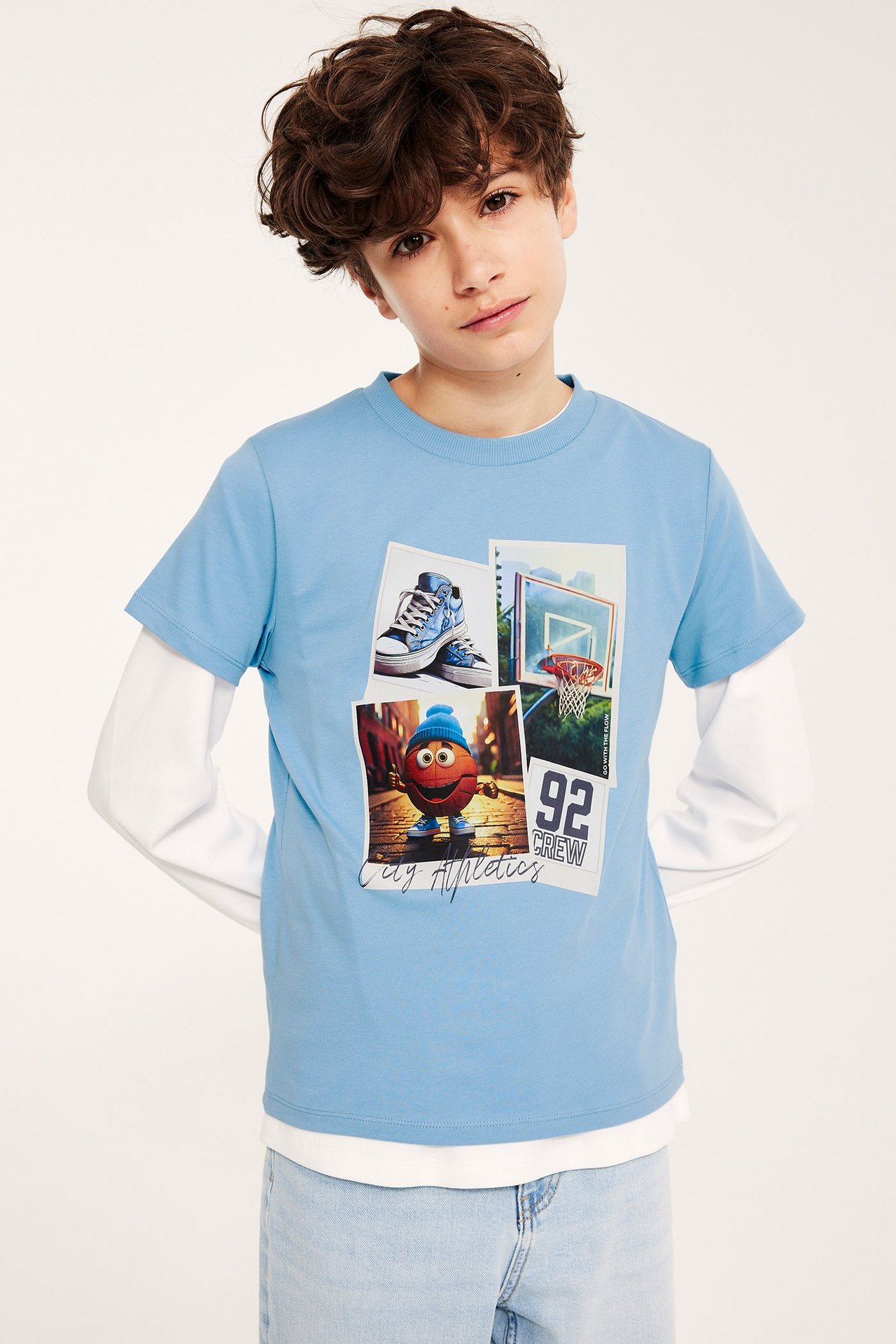 Edestä: Poika yllään vaaleansininen puuvillainen T-paita koripallopainatuksella valkoisen pitkähihaisen paidan päällä.