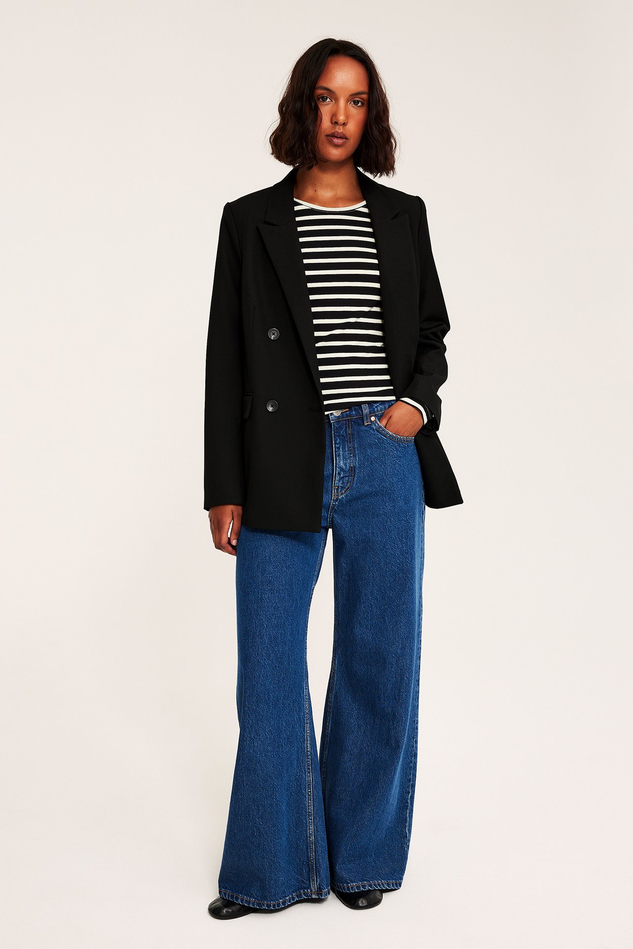 Front, voksen kvinne i åpen svart blazer over stripet topp og blå utsvungne jeans.