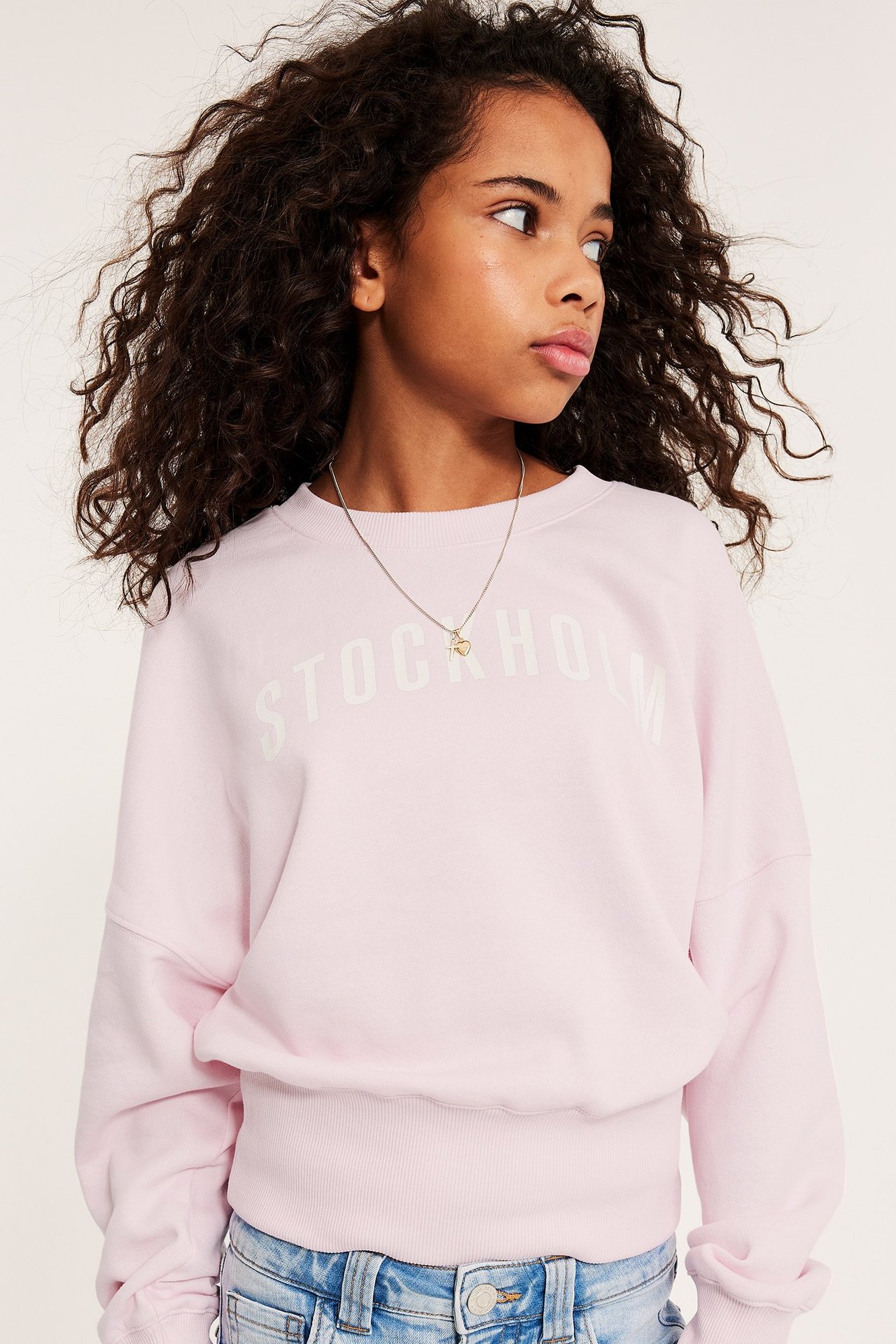 Forfra en jente i lys rosa genser med "STOCKHOLM"-tekst, lyseblå jeans og et sølvkjede.