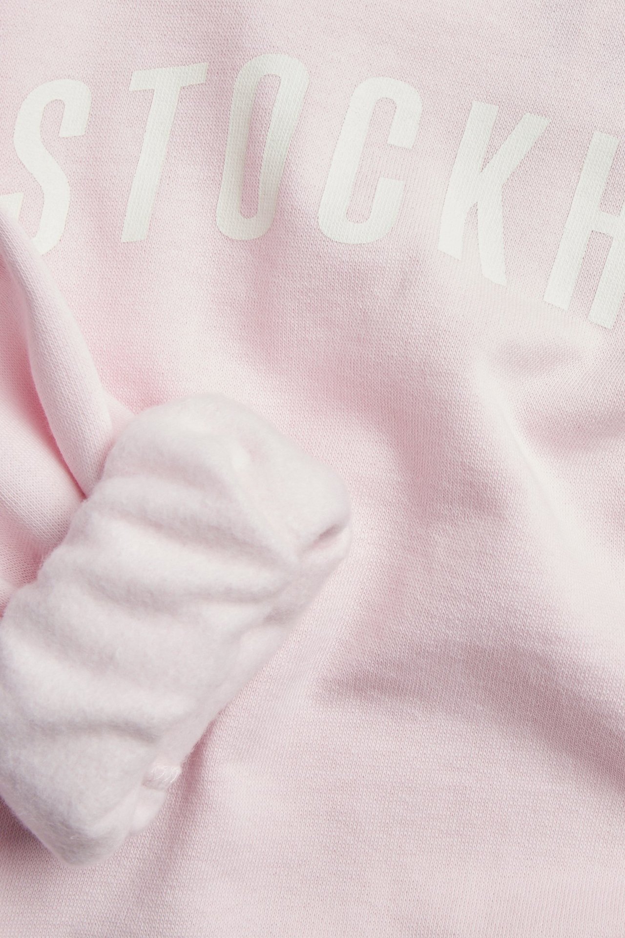 Nærbilde av en lys rosa collegegenser med hvit 'STOCKH'-tekst og ribbestrikket mansjett.