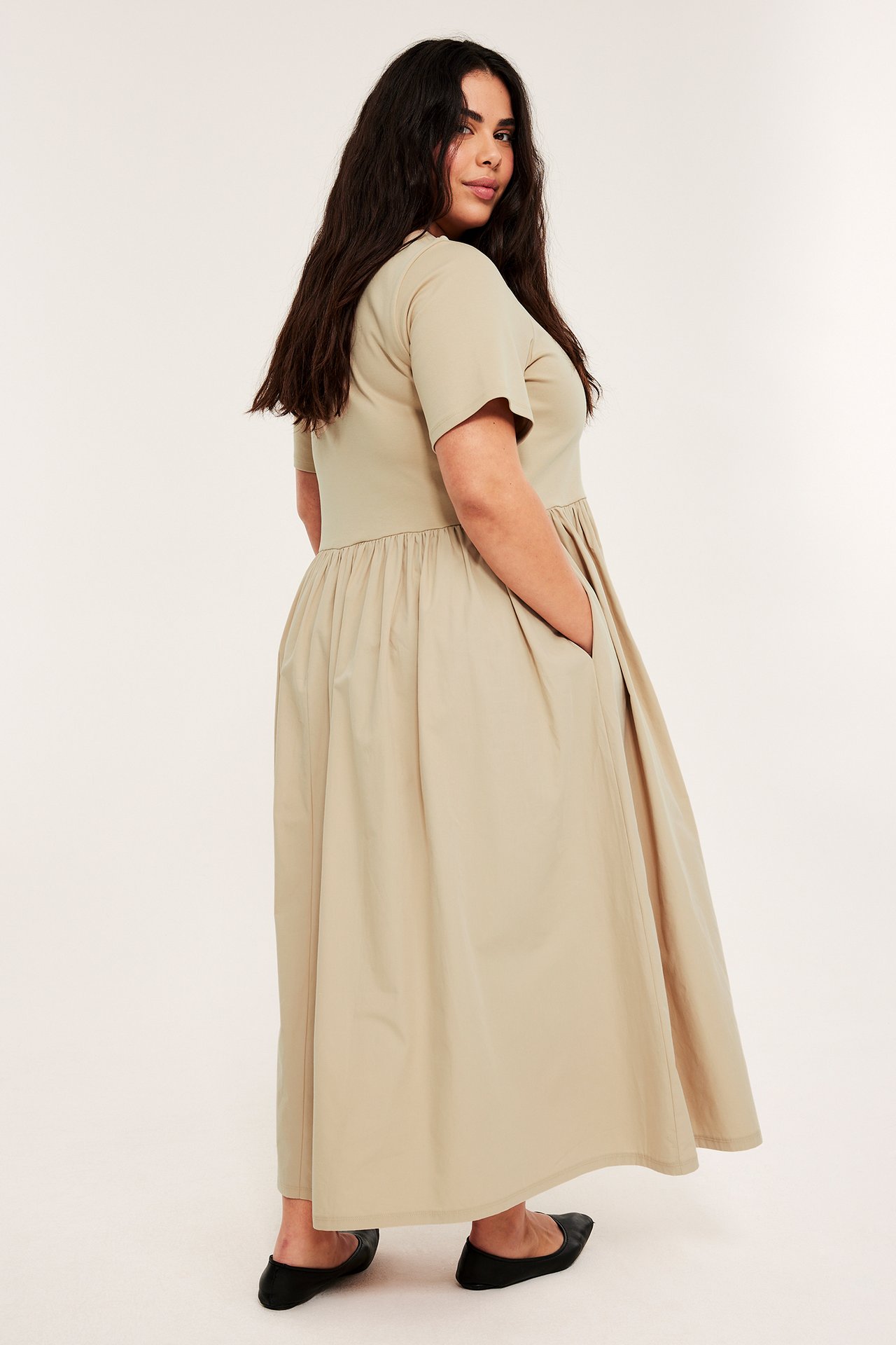 Nainen beigessä pitkässä mekossa, jossa lyhyet hihat, jerseymäinen yläosa, popliinihame ja mustat ballerinakengät.