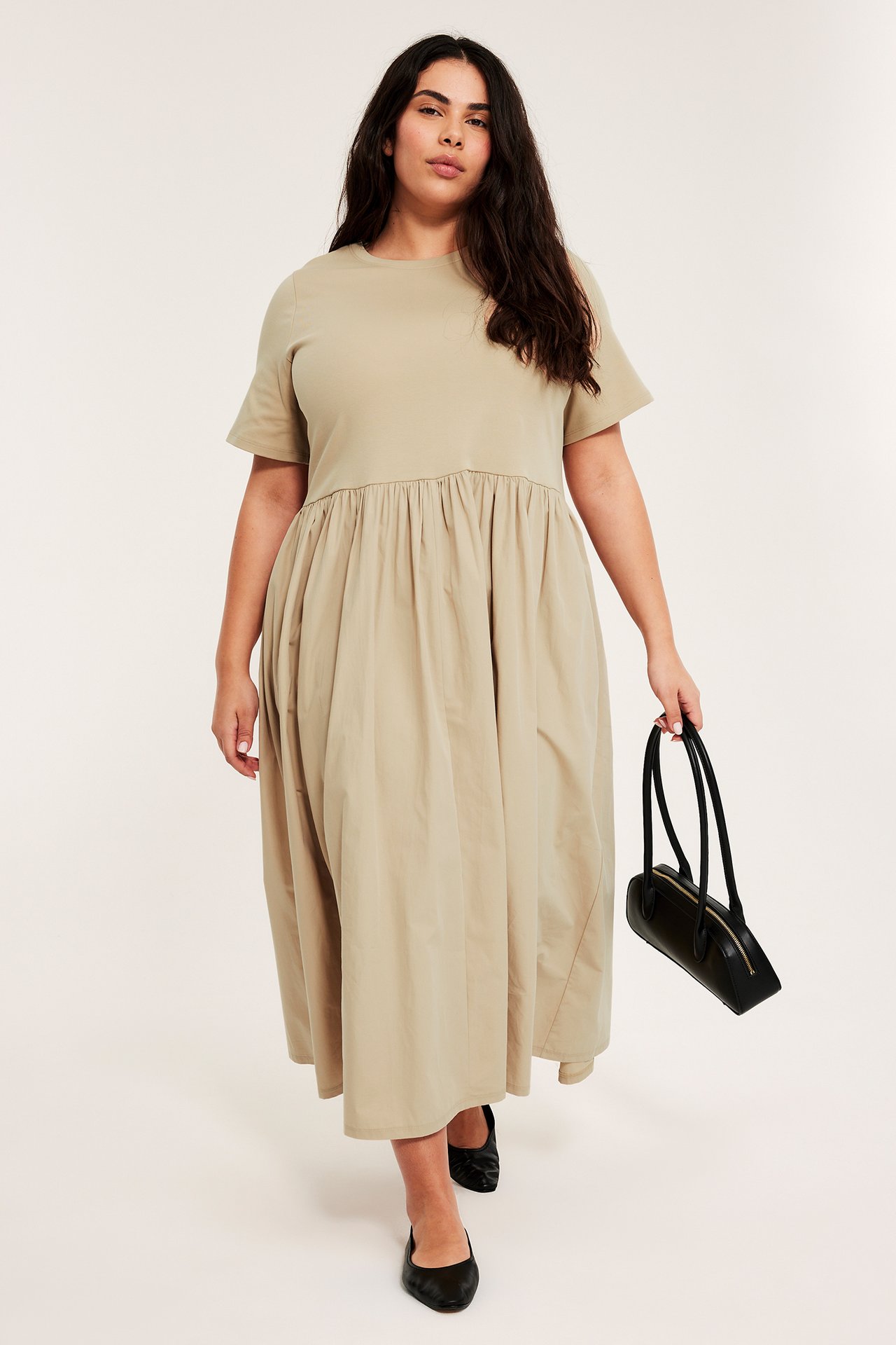 Frontbild: kvinna i beige kortärmad midiklänning, håller en svart handväska, bär svarta ballerinaskor.