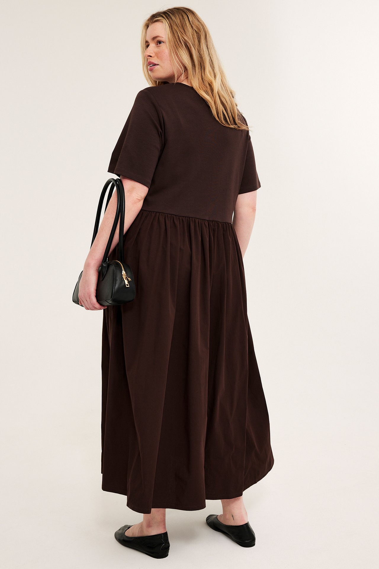 Widok 3/4 z tyłu: Kobieta w ciemnobrązowej sukience maxi, trzyma czarną torebkę, nosi czarne baleriny.