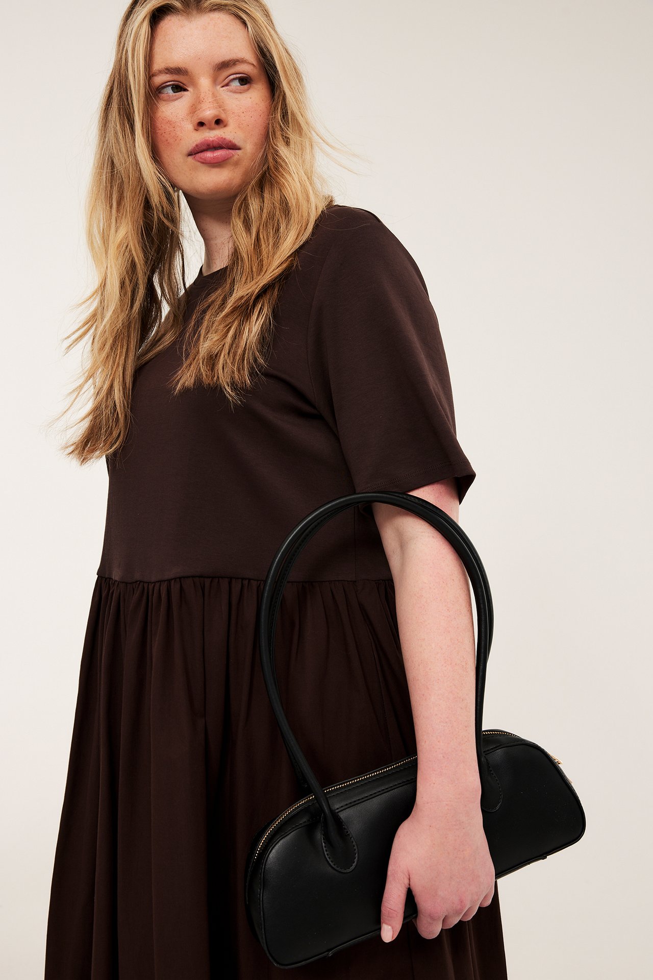 Framsida av kvinna i mörkbrun rundhalsad jerseyklänning med utställd poplinkjol, håller en svart handväska.